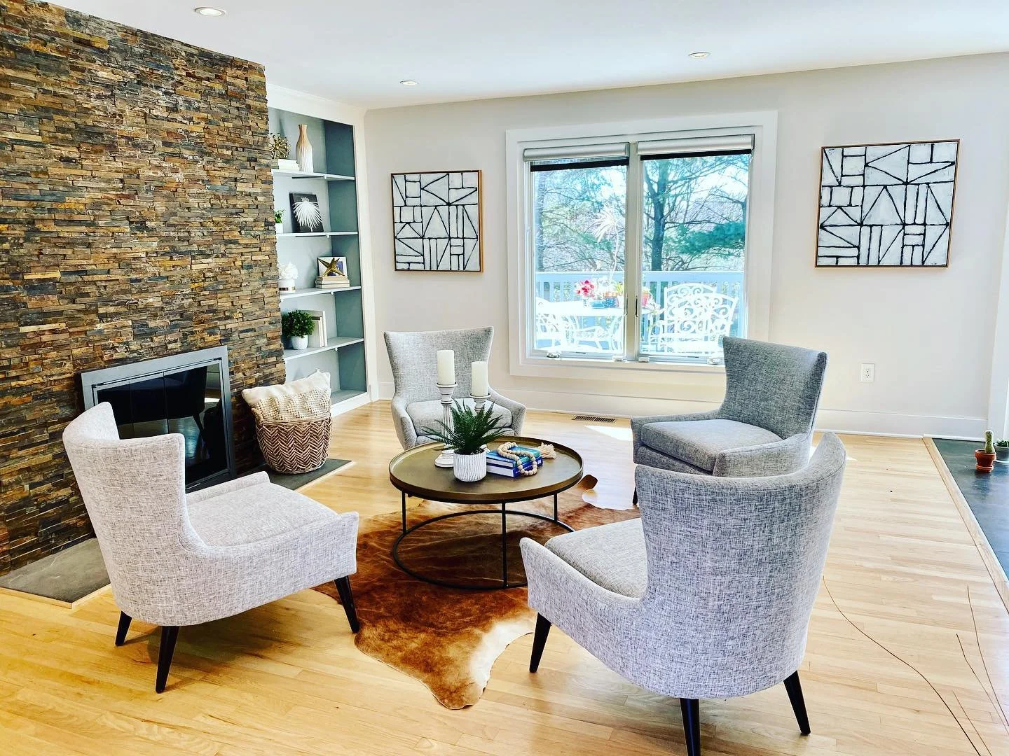 Living room with four gray armchairs around a round coffee table, a stone accent wall with a fireplace, a built-in shelving unit, and large window overlooking a balcony with outdoor furniture