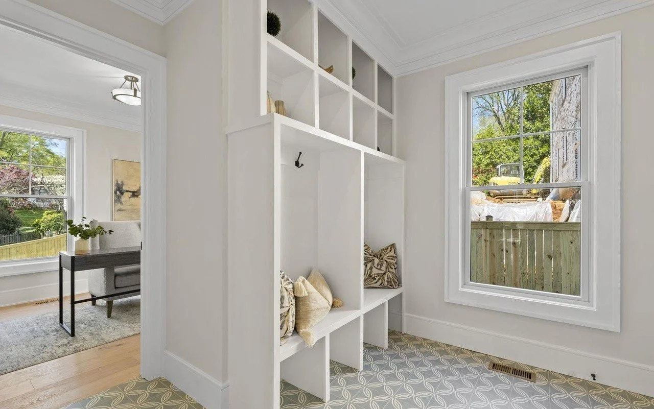 Interior of a bright room with large window showing trees outside, white shelving with decorative items, and cozy throw pillows on small seating area.