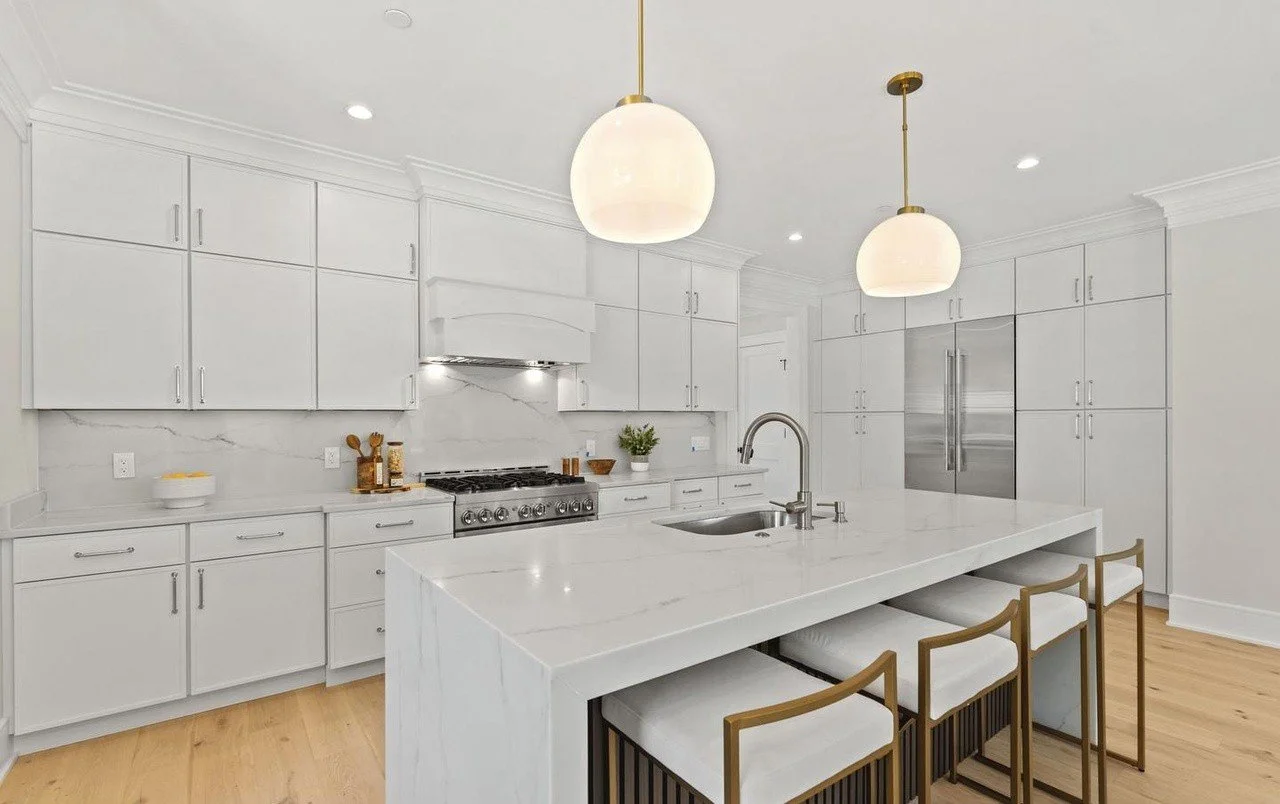 Modern white kitchen with marble countertops, stainless steel appliances, pendant lighting, and wooden barstools.