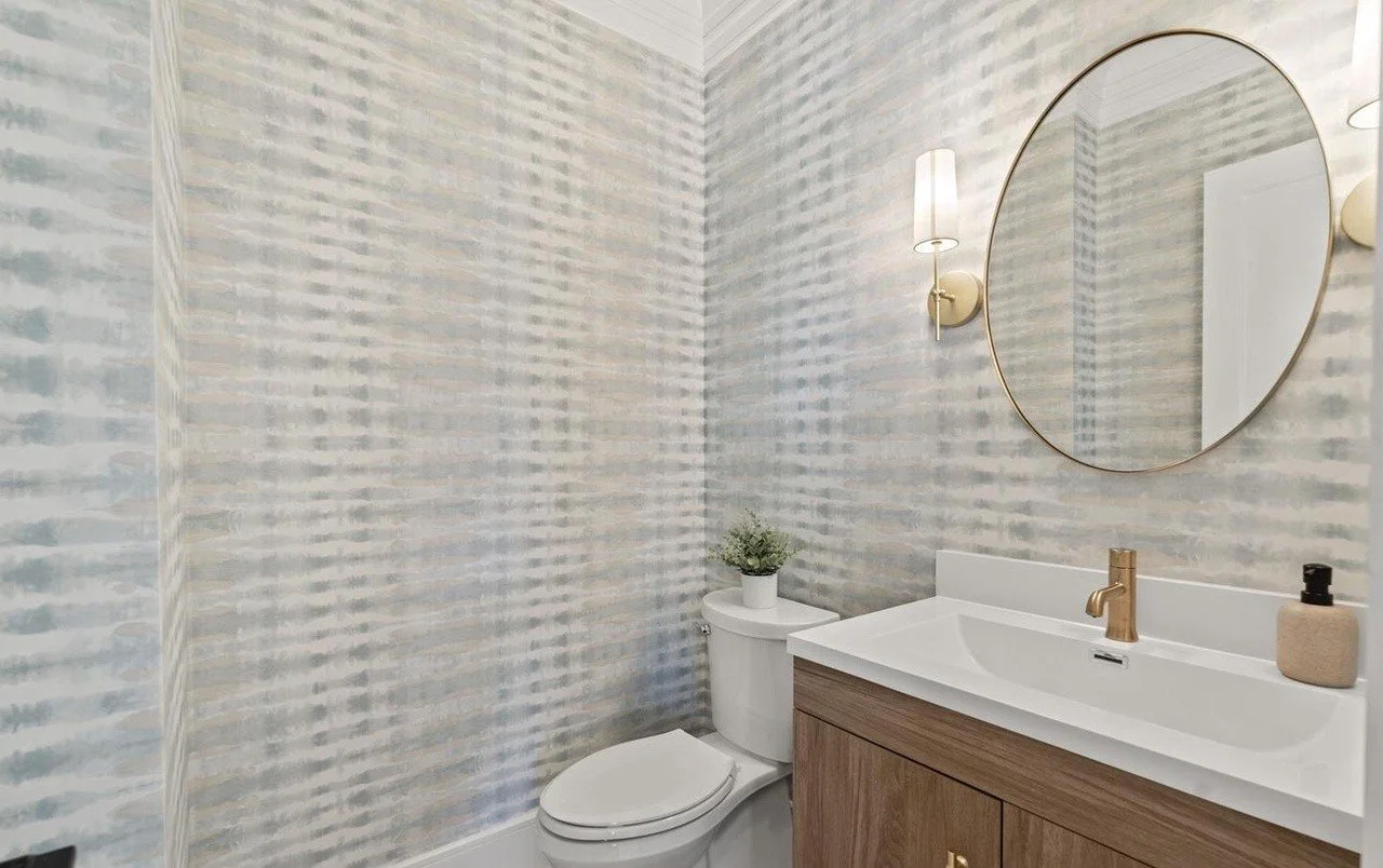 Modern bathroom with a toilet, wooden vanity with white countertop, round mirror, wall-mounted light fixtures, and a potted plant on the toilet tank.