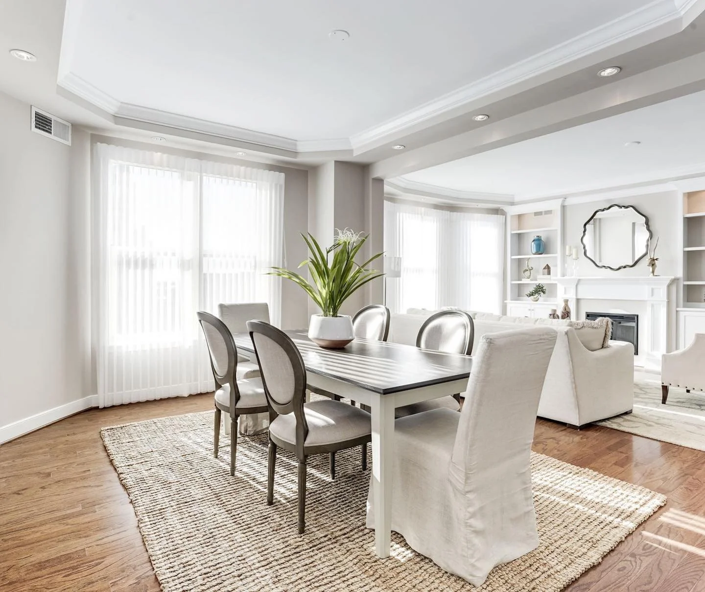 Bright, open living and dining room with white walls, large windows with sheer curtains, and hardwood floors. The dining area features a rectangular table with a potted plant centerpiece and six chairs, four with beige upholstery and two with slipcovers. The living space has a white sofa, armchair, area rug, and built-in white shelves with decorative items and a round mirror above the fireplace.