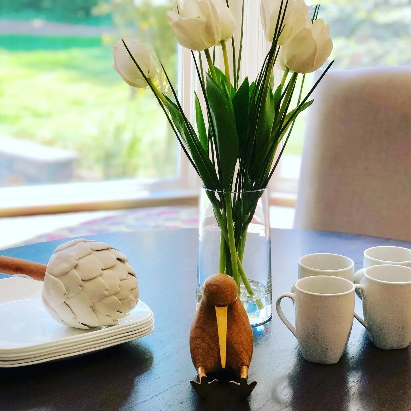 A vase of white tulips on a dining table with white coffee mugs, a wooden duck figurine, and a white pinecone decoration, with a window and greenery in the background.