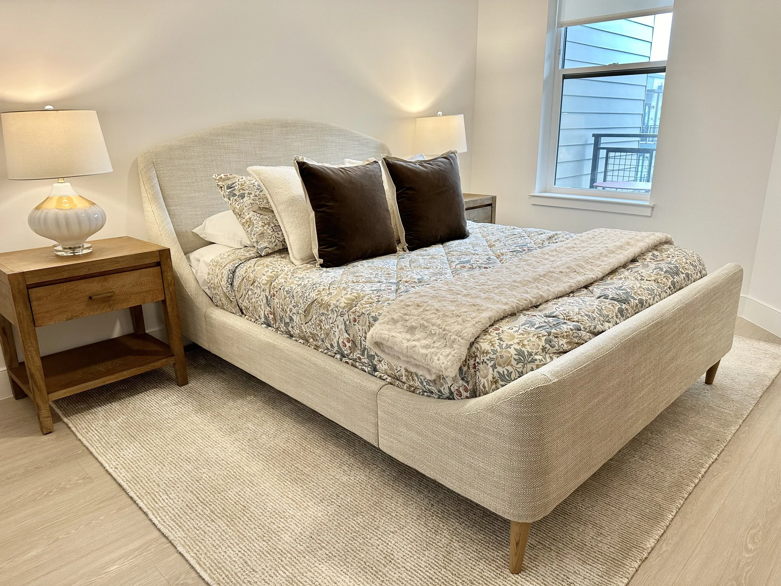 A bedroom with a beige upholstered bed, floral bedspread, and decorative pillows, flanked by wooden nightstands with lamps, and a window showing a neighboring building.