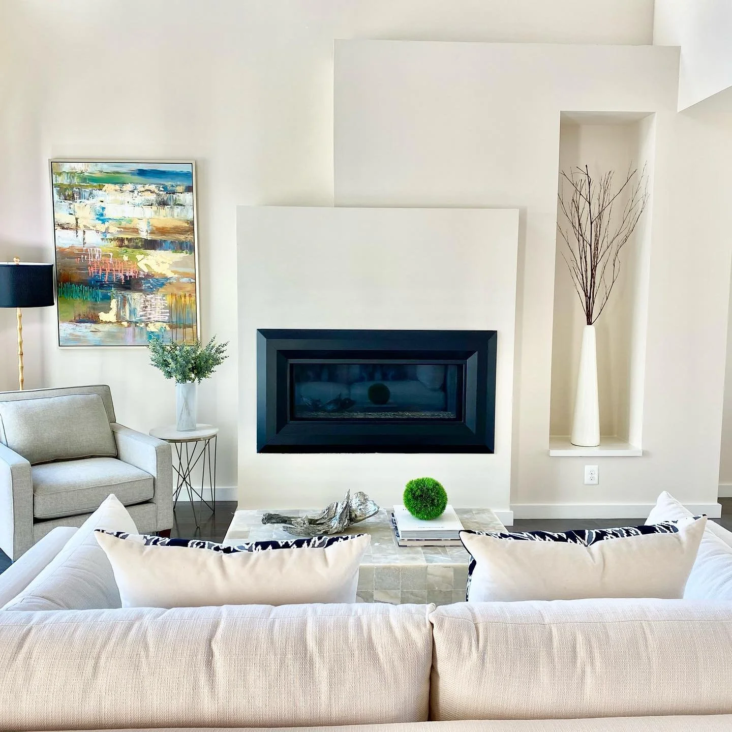 Modern living room with a fireplace, white sofa, gray armchair, abstract painting, tall vase with branches, and decorative items on a coffee table.
