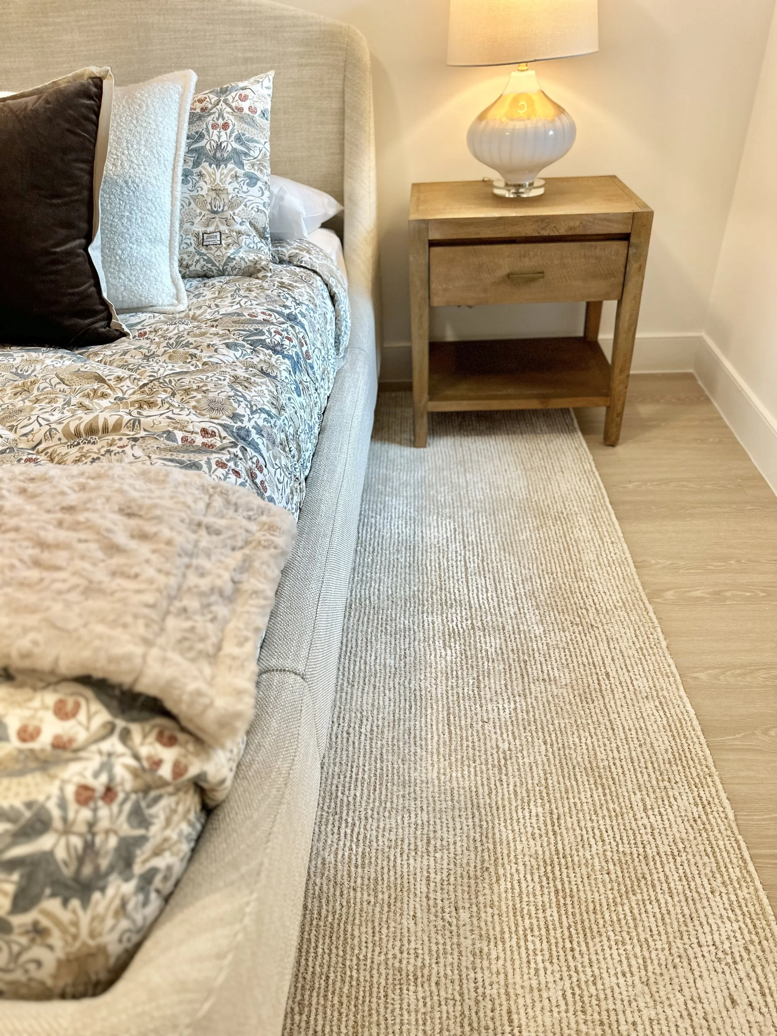 A beige upholstered bed with a floral comforter and various pillows, a wooden nightstand with a drawer and shelf, and a lamp on top, set on a beige striped rug with light hardwood flooring.