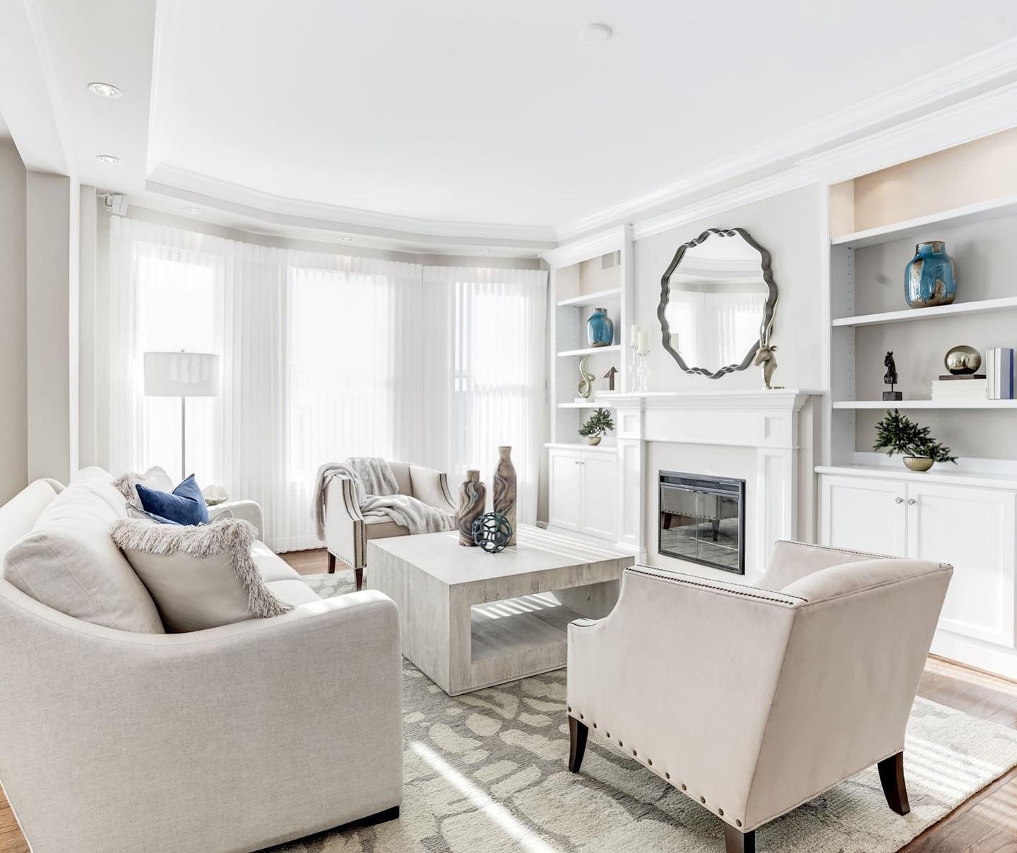 Bright living room with white sofas, armchair, marble coffee table, built-in white shelves, large windows with sheer curtains, fireplace, decorative vases, mirror, and minimal décor.