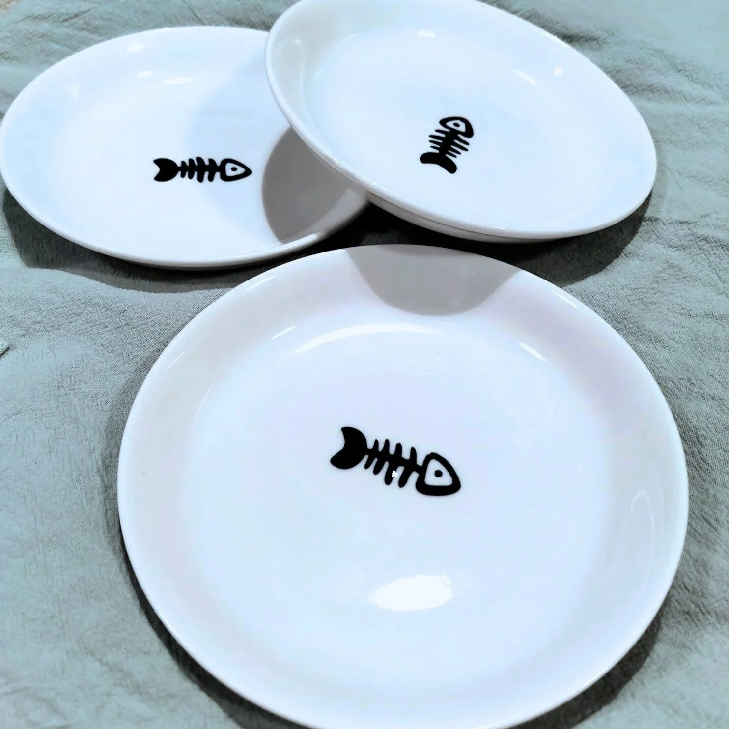 three white shallow bowls with black stick drawing of fish bones used for cat bowls