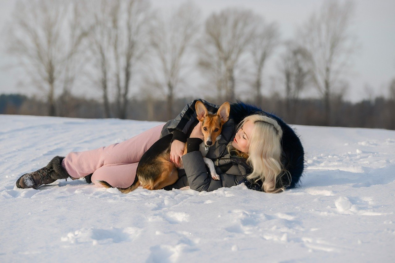Young woman lying in the snow with a dog cuddled in her arms