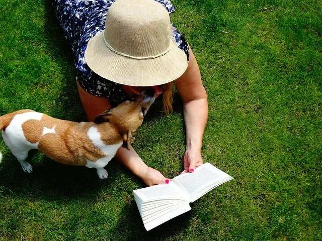 woman lying on grass while reading with small dog licking her face