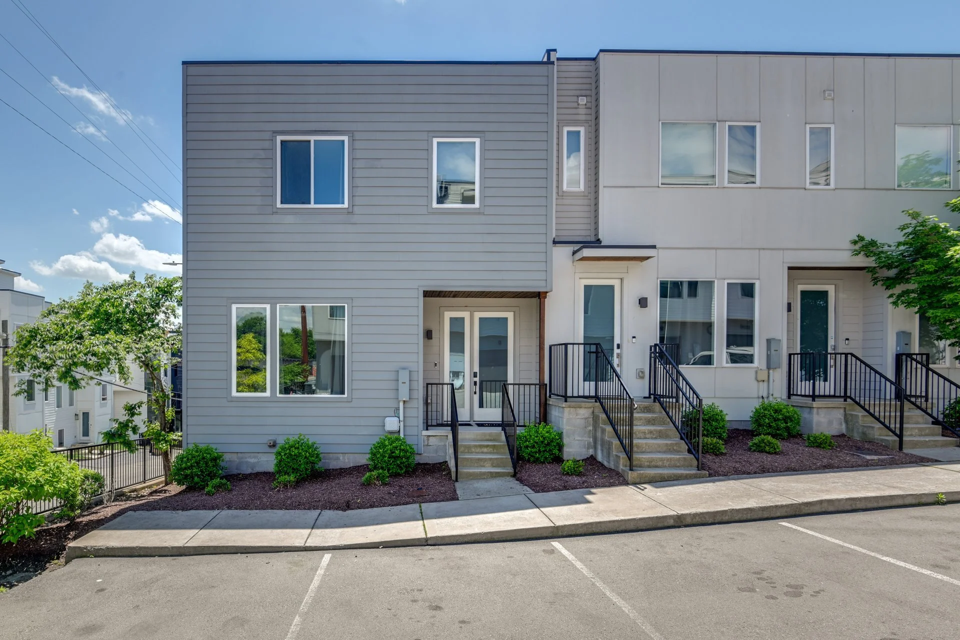 Modern multi-story residential townhouses with steps, landscaped bushes, black railings, and parking spaces in front.