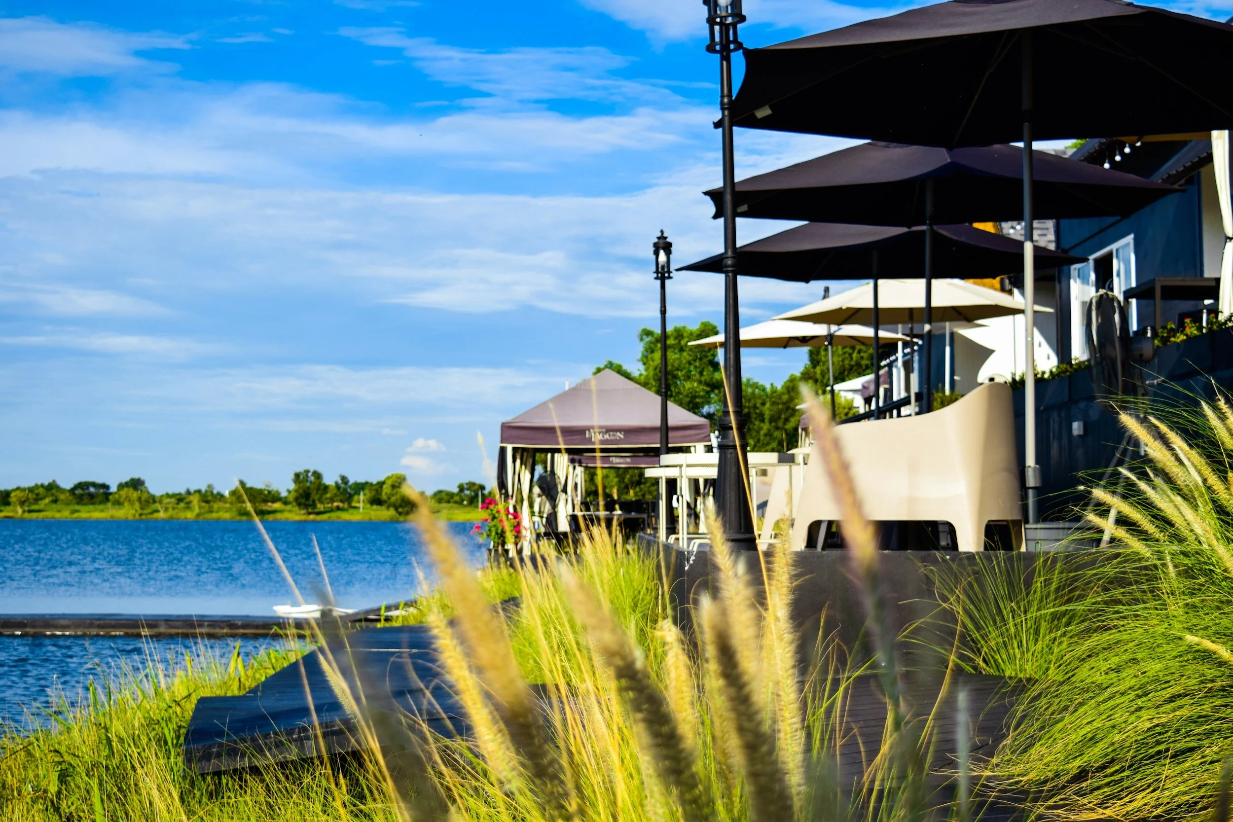 Outdoor lakeside restaurant with umbrellas, outdoor seating, and greenery, under a blue sky.