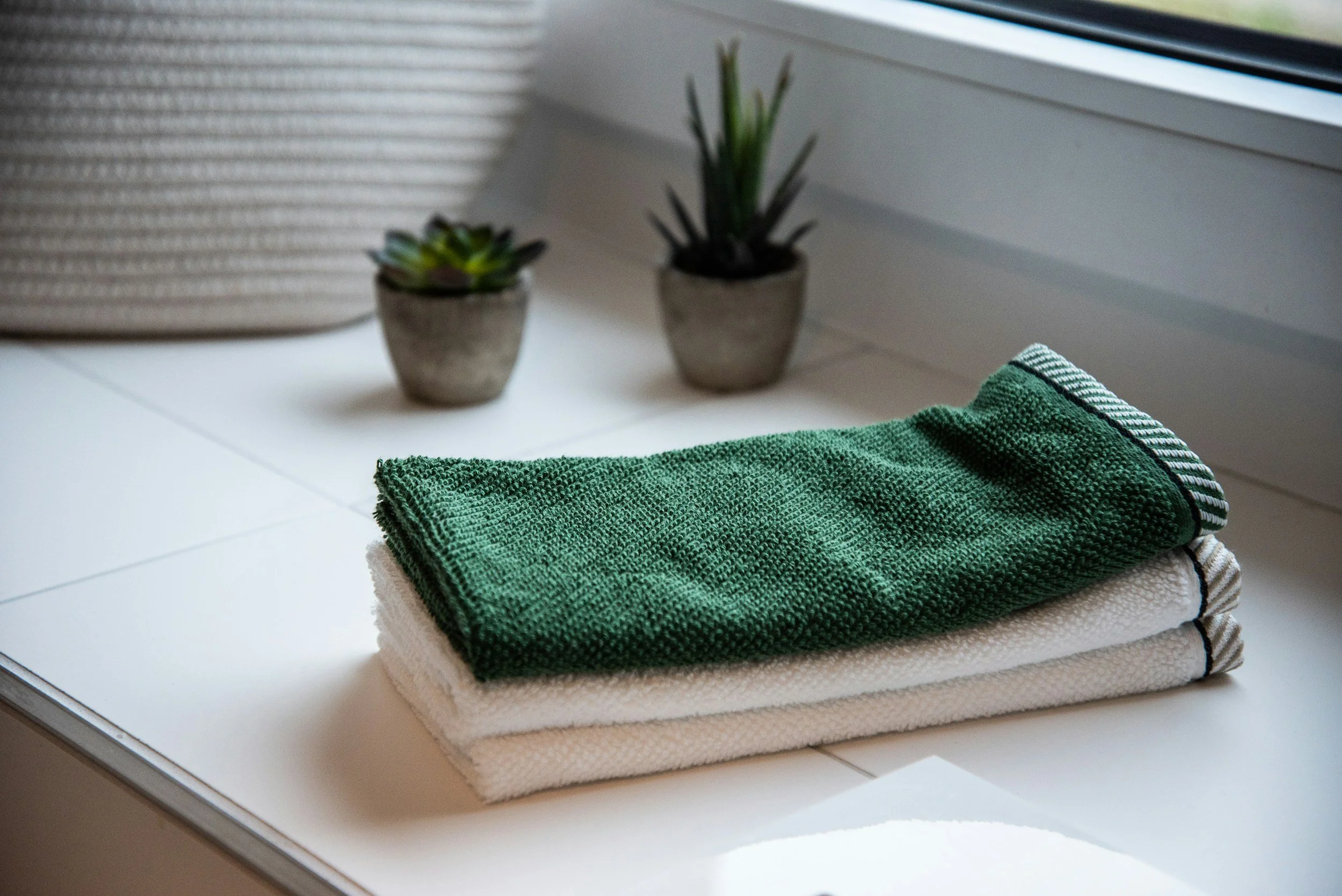 Stacked folded white and green kitchen towels on a white surface near a window, with two small potted succulents in the background.
