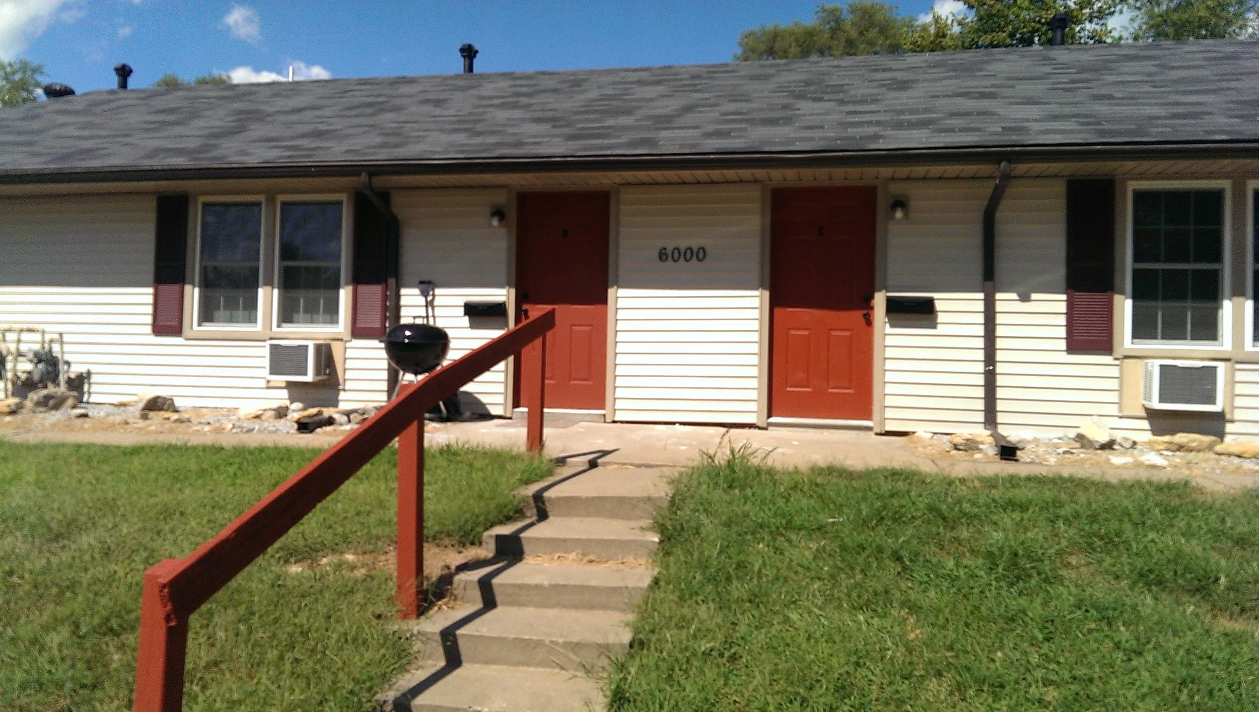 Residential building with two apartment doors, each painted red, and a set of concrete stairs with a red railing leading up to the entrances. The building has white siding with black shutters and window air conditioning units.