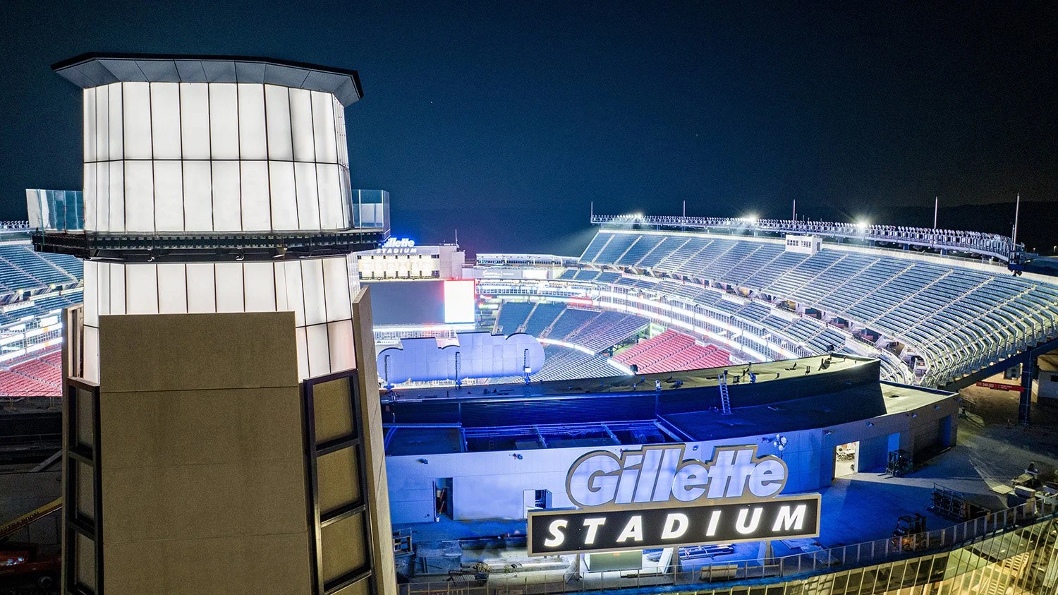 Night view of Gillette Stadium with empty seats and bright lighting, featuring the stadium's sign and a large structure in the foreground.