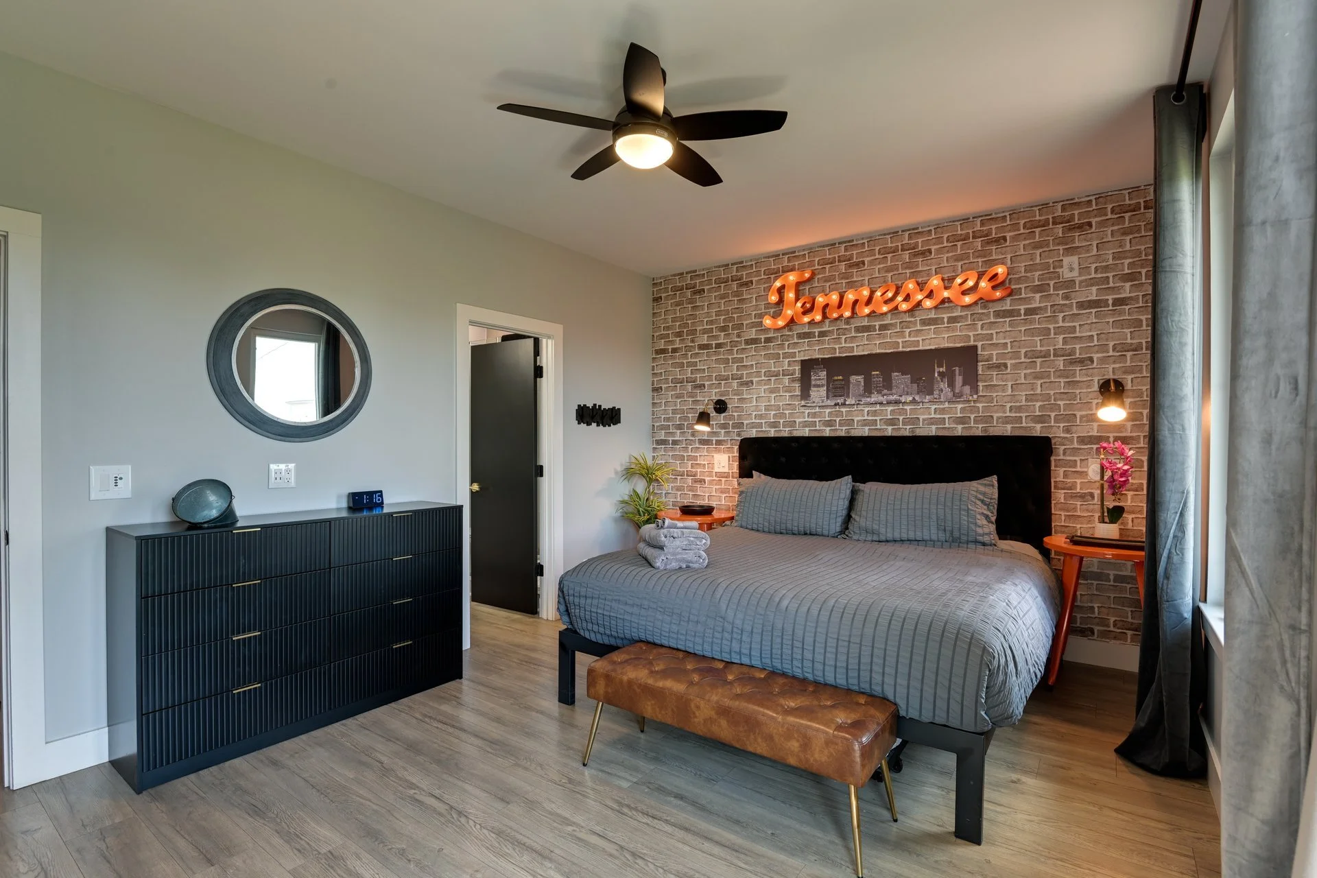 Modern bedroom with a bed, black headboard, brick accent wall, neon sign reading 'Tennessee,' side table with a plant, and black side wall lights. There is a black dresser with a mirror above, a ceiling fan, and large window with curtains.