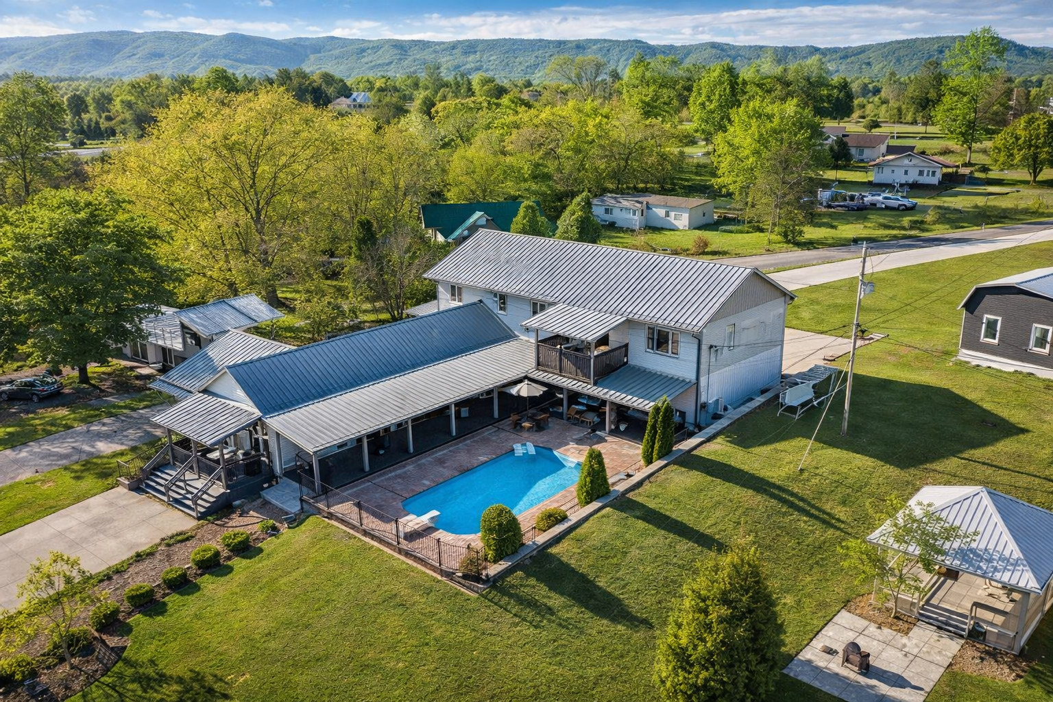 Aerial view of a house with a backyard, swimming pool, and covered patio, surrounded by lush green trees and open fields with mountains in the distance.