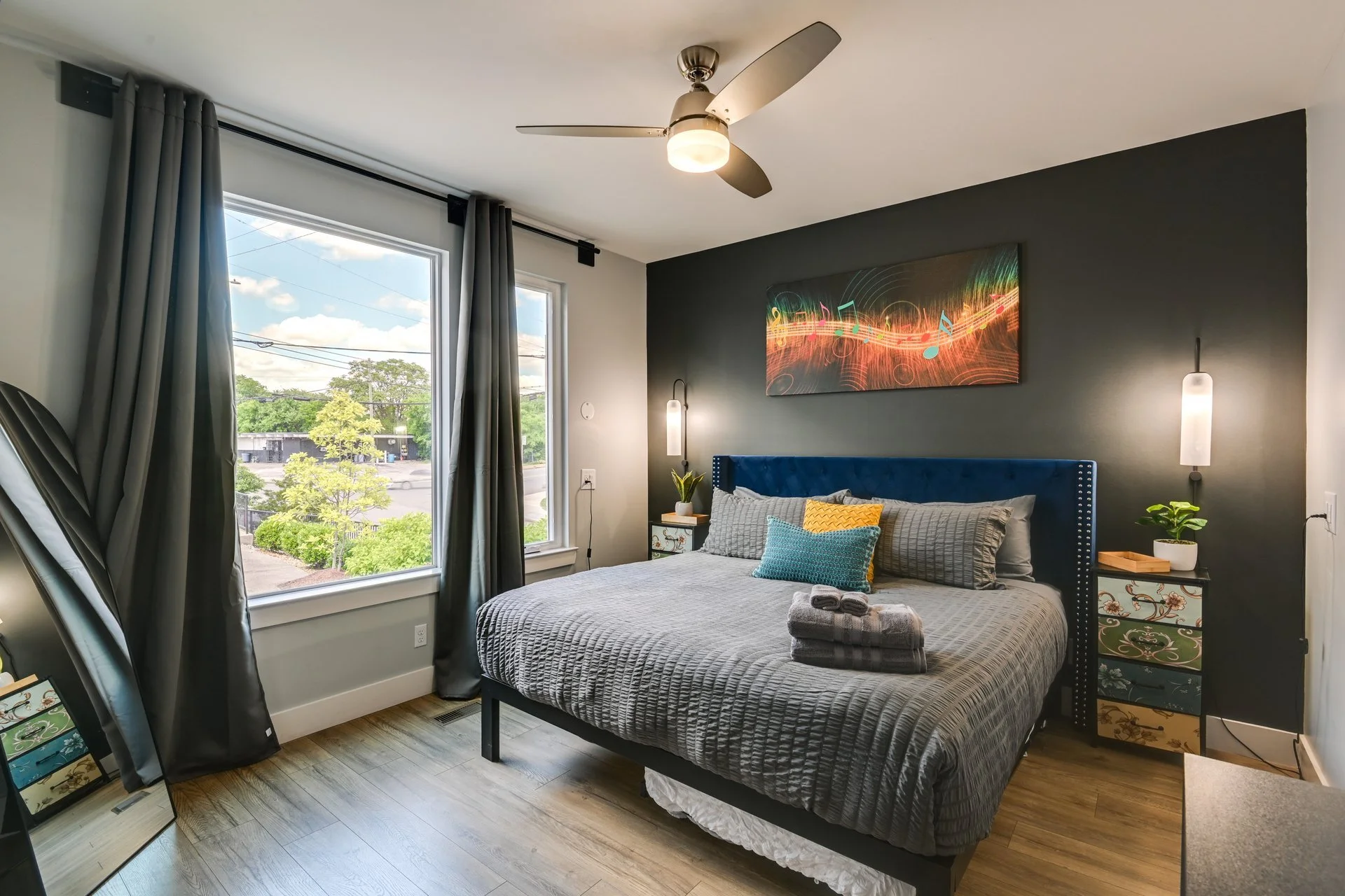 Modern bedroom with large window, gray curtains, dark accent wall, and ceiling fan; bed has a blue headboard, colorful pillows, and a rolled towel set on top; nightstands with decorative drawers, plants, and lighting; abstract artwork with musical no