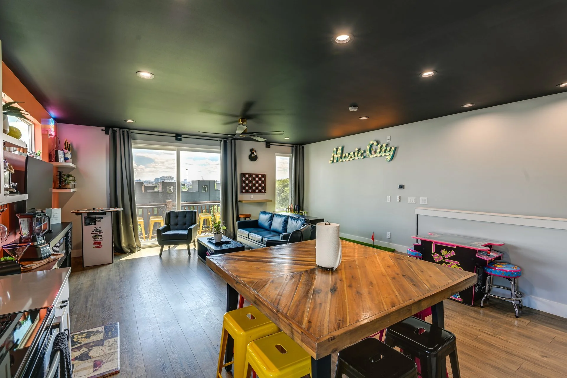 Living room with wooden dining table, black leather sofas, large window with balcony, family entertainment area, and a 'Music City' sign on the wall.