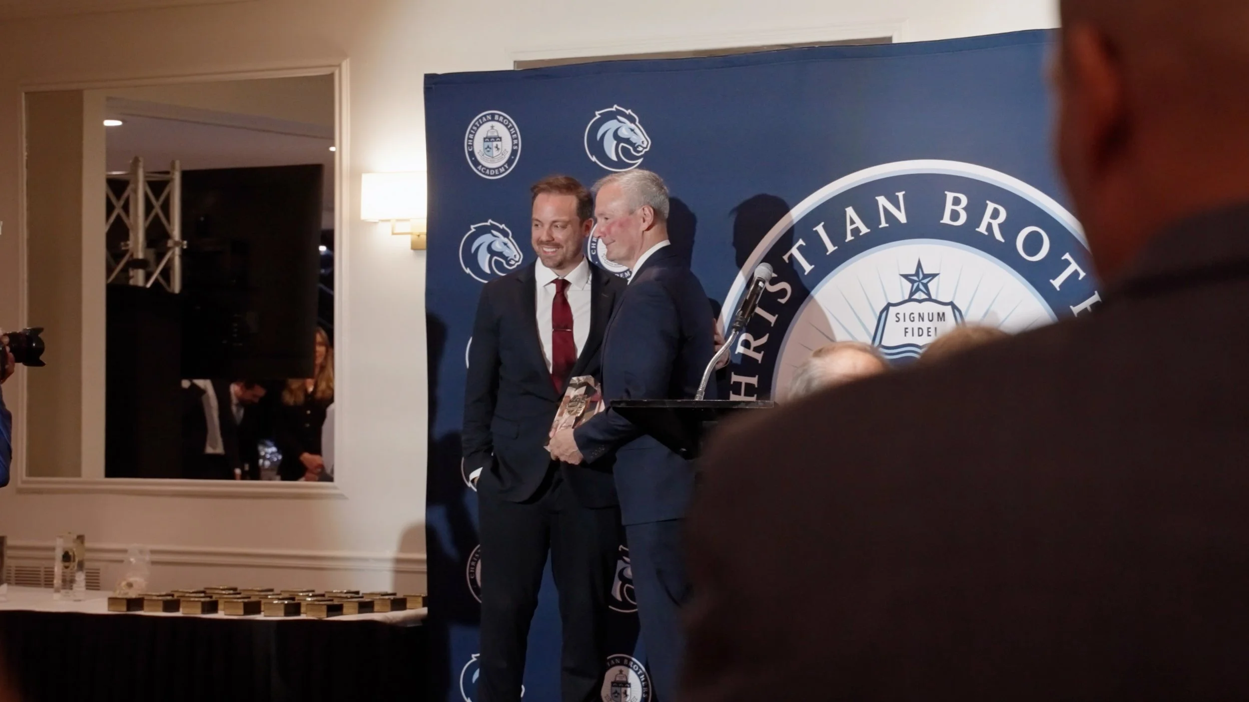 Two men in suits, one holding a plaque, smile on stage at a Christian Brothers Academy event with a blue backdrop featuring the academy logo.