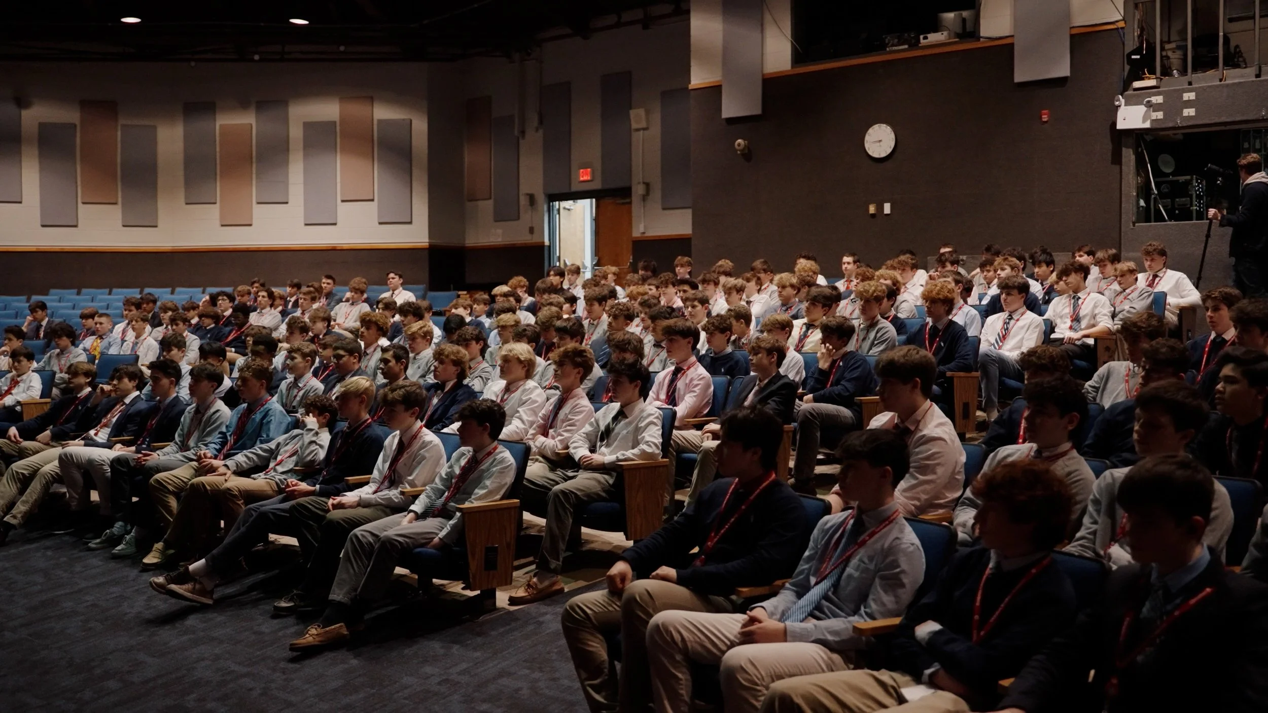 A large group of young boys seated in an auditorium or conference room, dressed in formal attire with lanyards around their necks, attentively listening to a presentation or speaker.