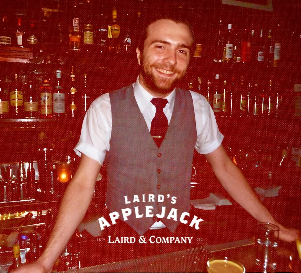 A smiling bartender with a beard and mustache in a gray vest, white shirt, and red tie, standing behind a bar with liquor bottles in the background. The bar displays the text "LAIRD'S APPLEJACK LAIRD & COMPANY."