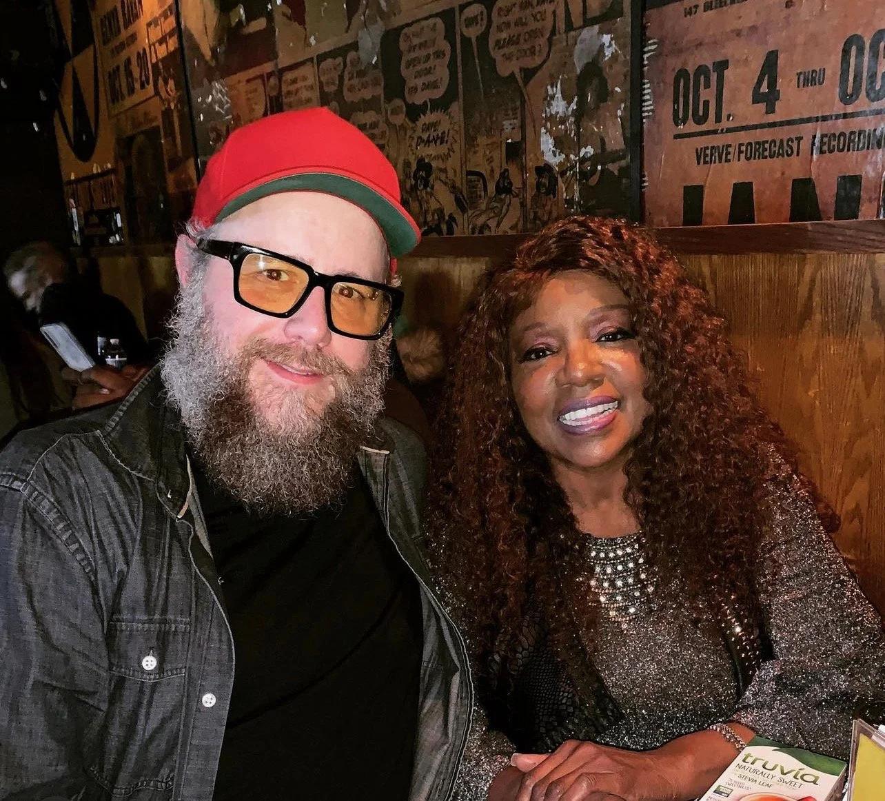 A man with a beard, glasses, and a red cap taking a selfie with a woman with curly hair and makeup at a restaurant.