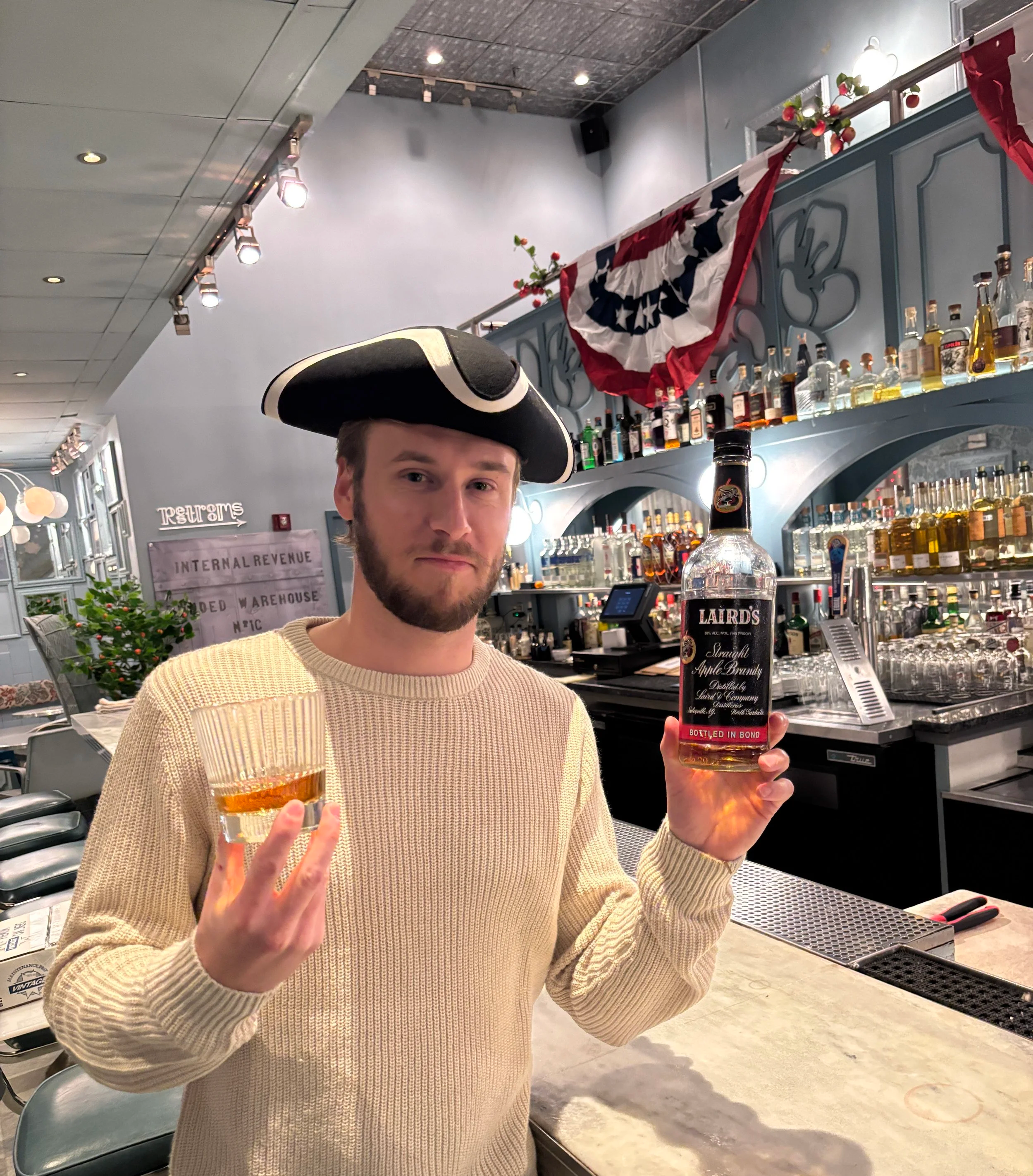 A man dressed as a pirate holding a glass of amber liquor in one hand and a bottle of Laird's apple brandy in the other, standing at a bar with bottles of alcohol in the background, decorated with patriotic bunting and a small Christmas tree.