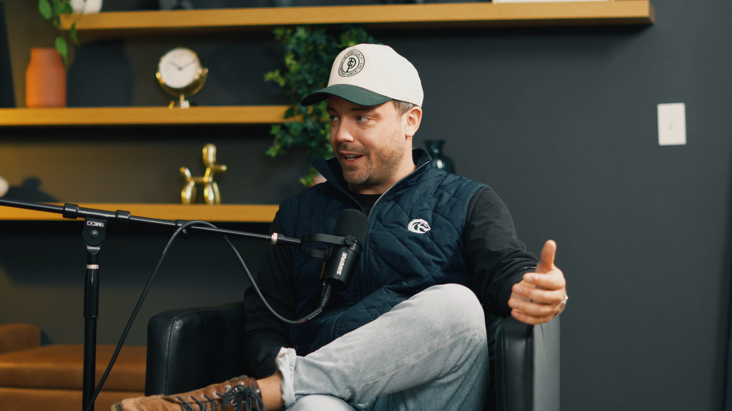 A man wearing a white cap, a black jacket, and light-colored pants sitting in a black leather chair, speaking into a microphone during a podcast or interview in a room with dark walls and wooden shelves decorated with plants, a clock, and a gold dog figurine.