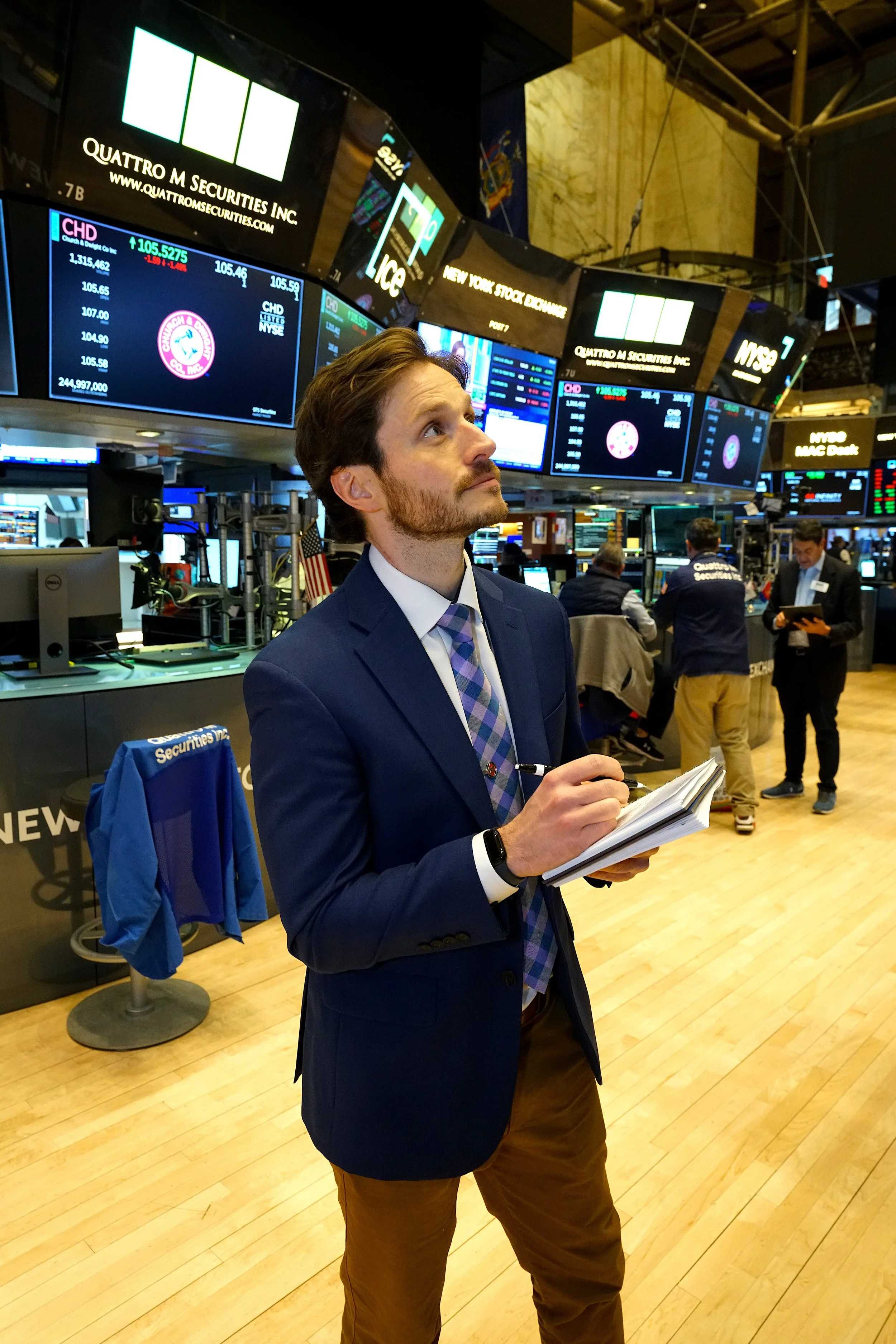 A man standing on the trading floor of the New York Stock Exchange, holding a notebook and pen, with multiple electronic stock market displays above him.