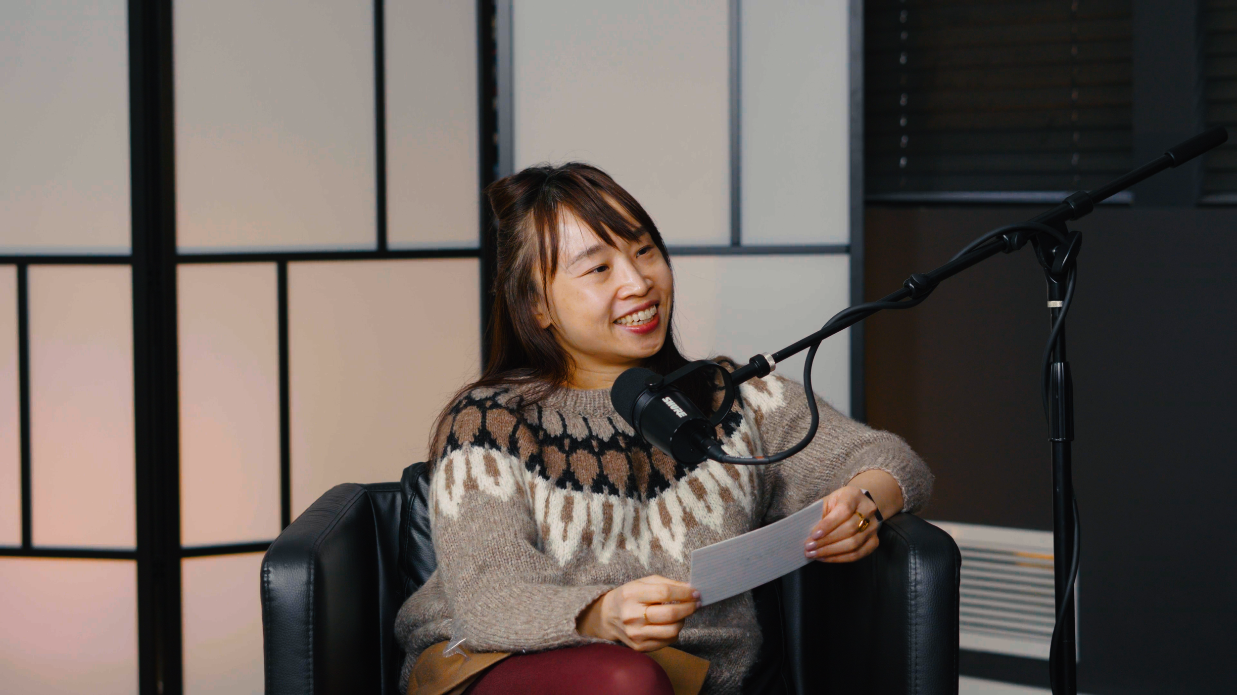 A woman with long dark hair, wearing a patterned sweater, sitting on a black leather chair, holding a paper, speaking into a studio microphone, smiling during a recording session.