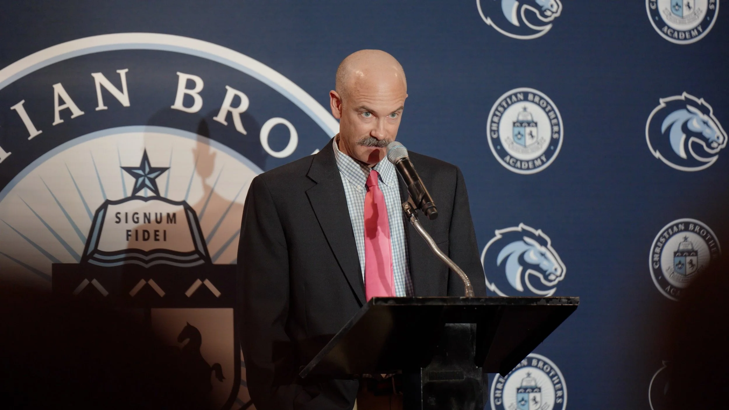 A man with a bald head and mustache in a suit and pink tie stands at a podium, speaking into a microphone during a formal event. The background features logos of Christian Brothers Academy and a large emblem with the Latin phrase 'Signum Fidei'.