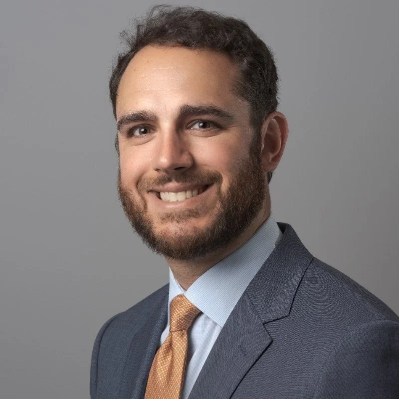 A portrait of a smiling man with dark curly hair and a beard, wearing a dark suit, light blue shirt, and orange tie, against a plain gray background.