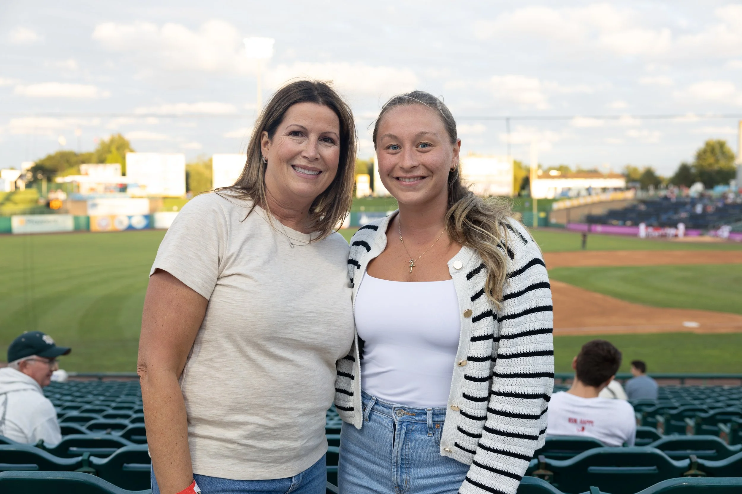 Two women standing together at a baseball stadium, smiling at the camera with the field and spectators in the background.