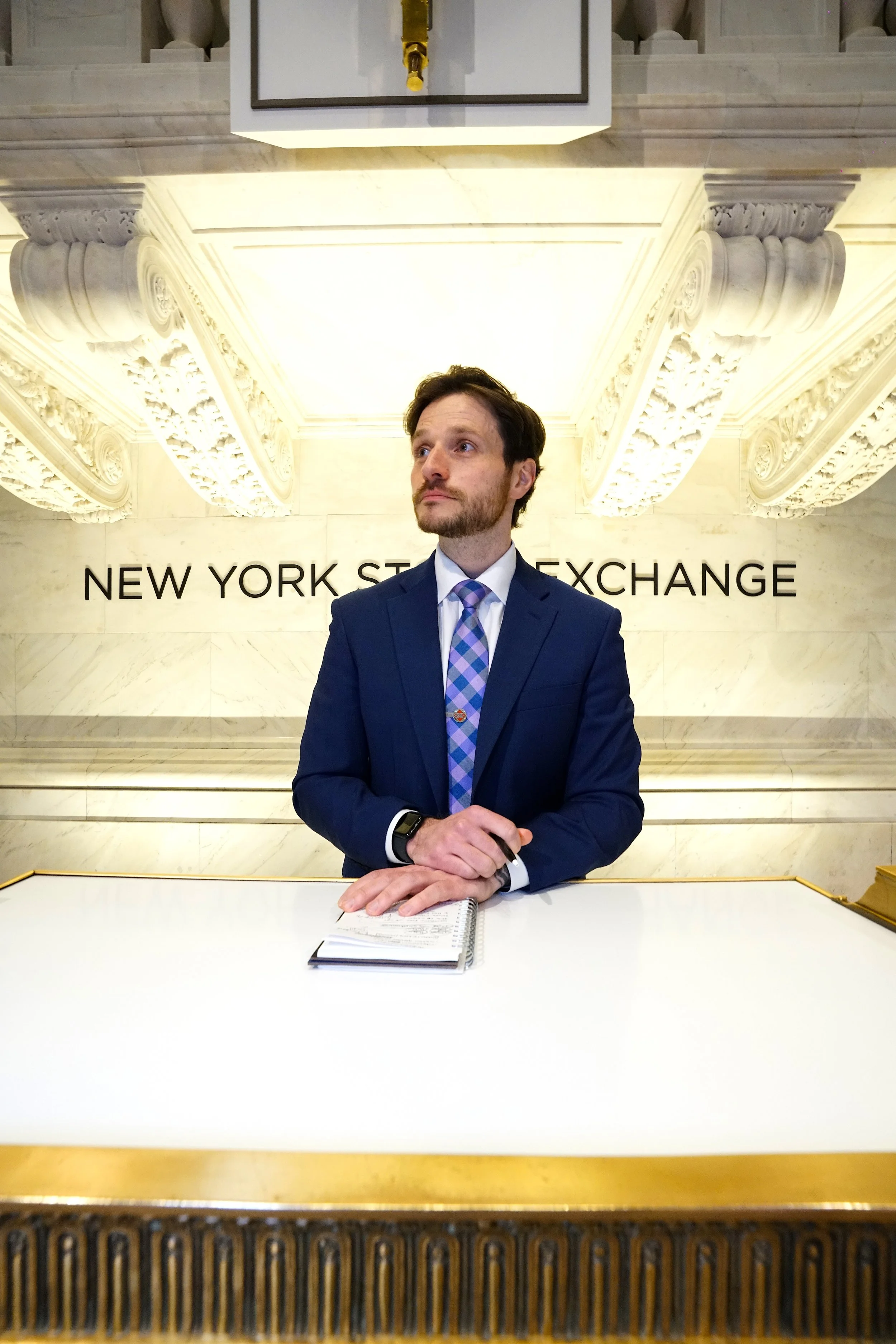 A man in a blue suit standing at a white and gold reception desk at the New York Stock Exchange. He has a notebook in front of him and is looking slightly to the side. The background shows ornate white architectural details and the NYSE sign.