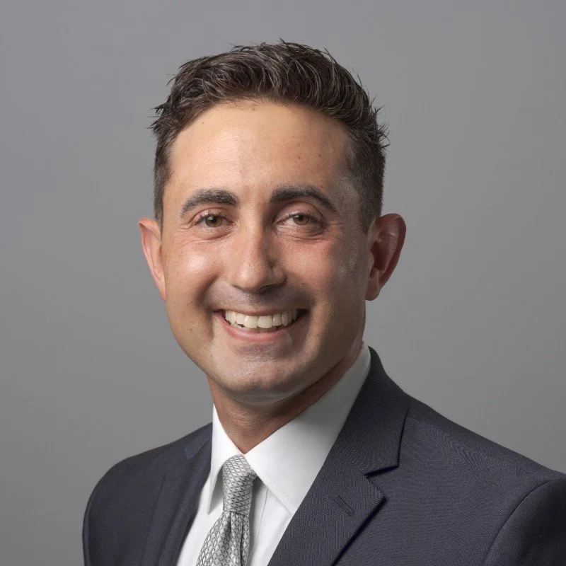 Professional headshot of a man in a dark suit, white shirt, and patterned tie, smiling against a neutral gray background.