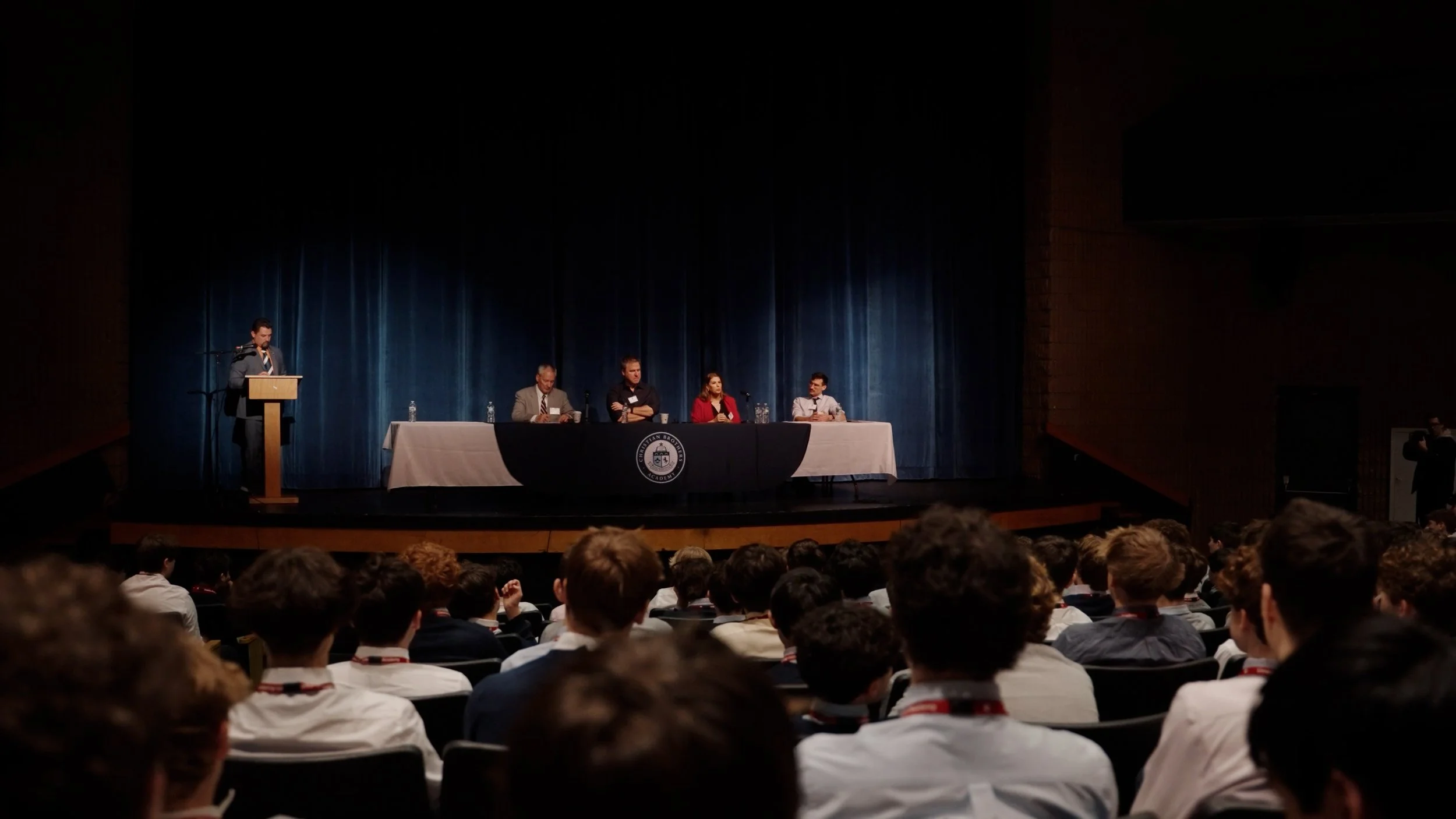 A panel discussion on stage with five people sitting at a long table, and one person standing at a podium speaking to an audience in an auditorium with a blue curtain background.