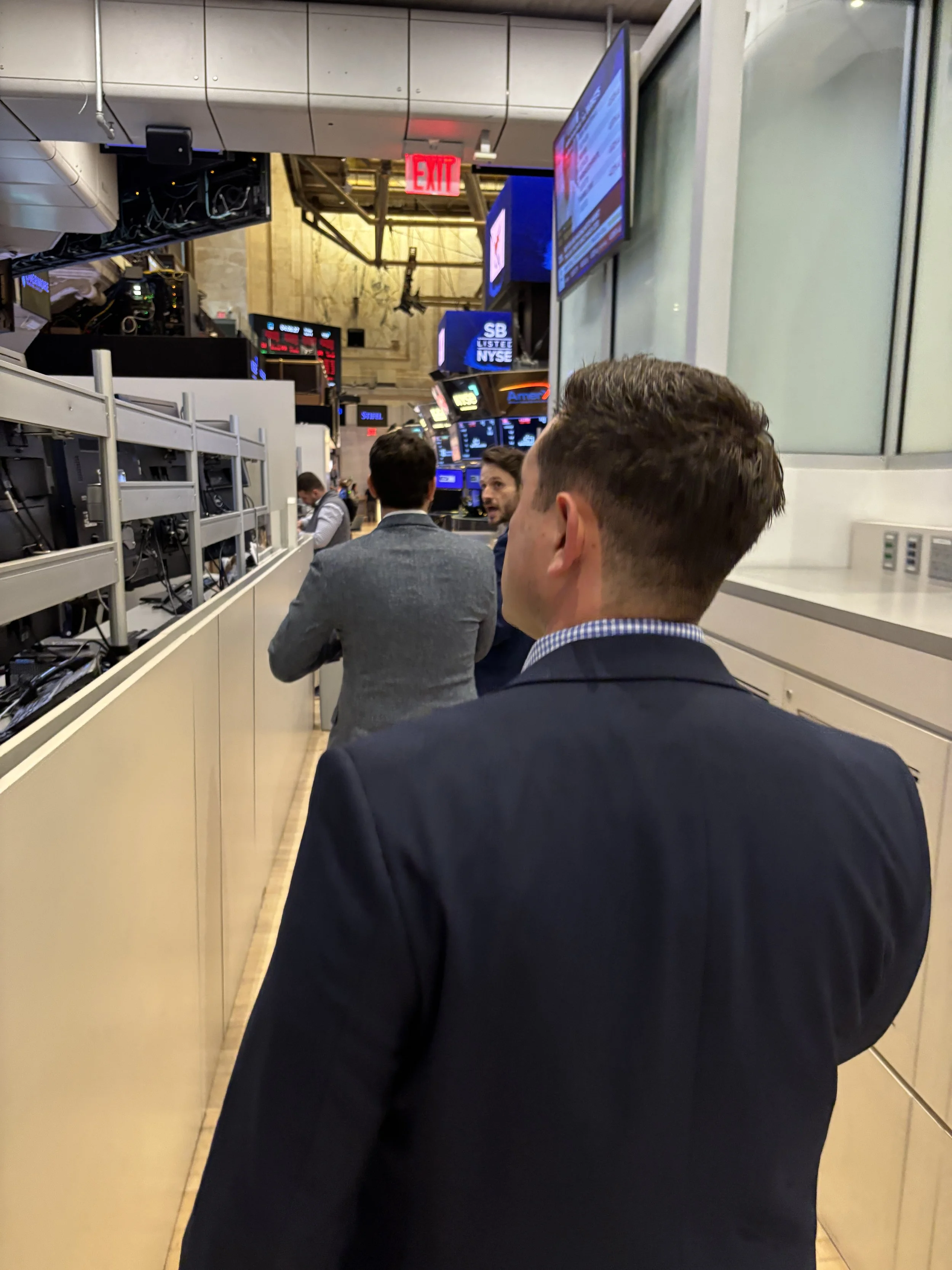 People standing in line on the trading floor of the New York Stock Exchange, with multiple electronic stock ticker displays and monitors showing financial information above them.