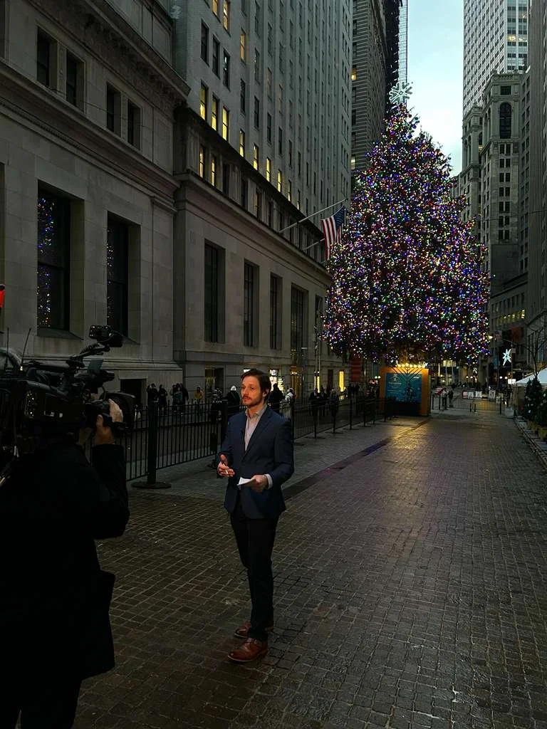 A man in a suit being interviewed by a cameraman on a city street with tall buildings, a decorated Christmas tree with colorful lights, and an American flag.
