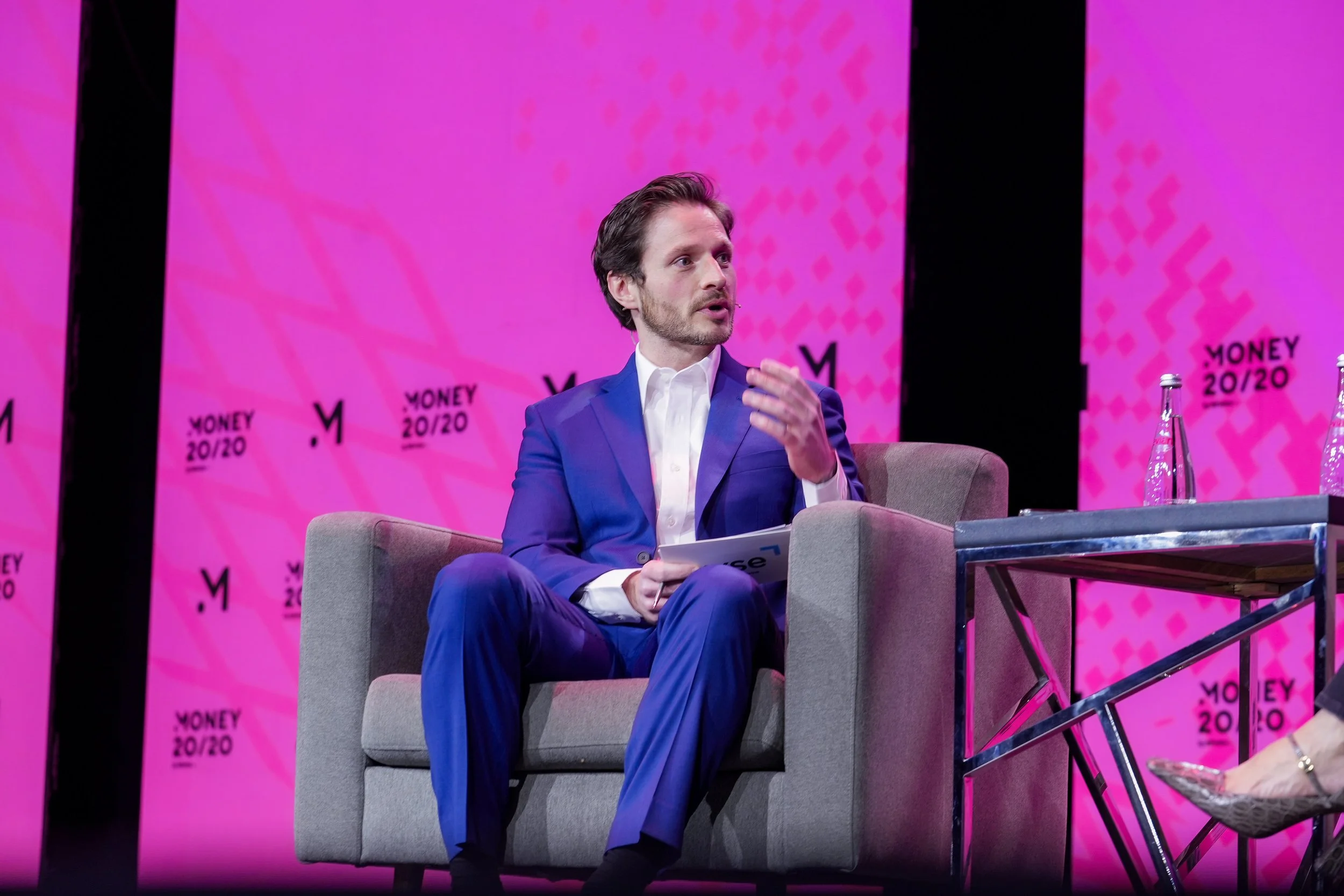 A man in a blue suit sitting on a gray armchair on stage, gesturing with his hand during a panel discussion at the Money 20/20 conference. The background features a large pink screen with the event name and logo.