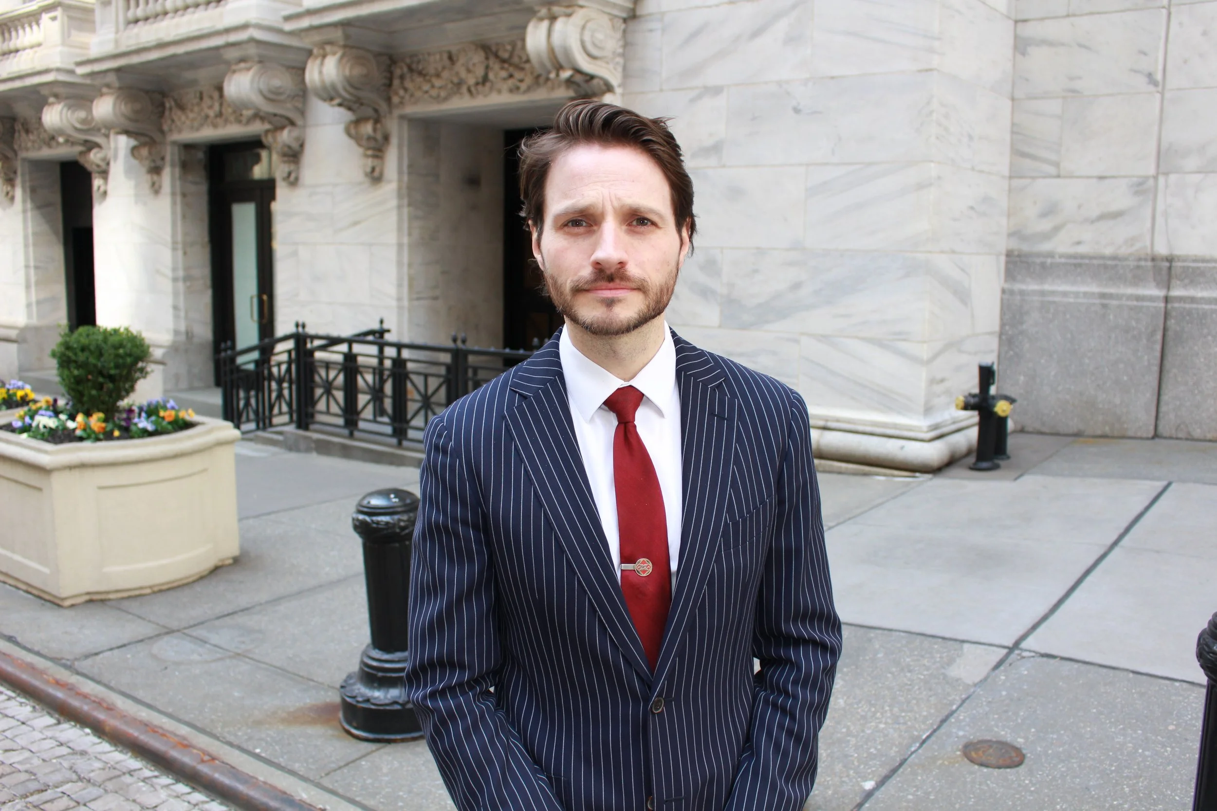 A man in a navy blue pinstripe suit with a white shirt and red tie standing on a city sidewalk in front of a beige stone building, with a planter and black bollards nearby.