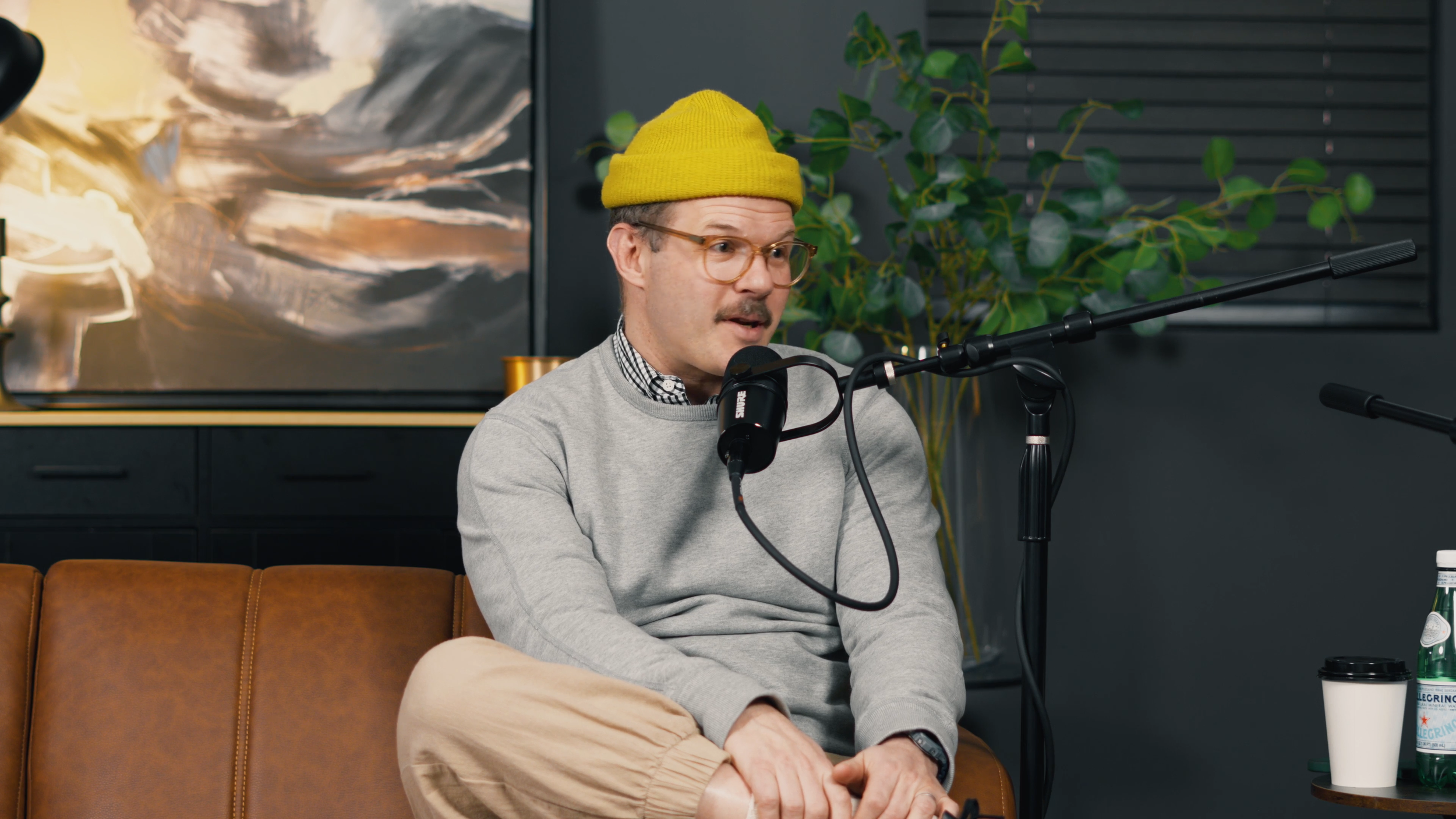 A man wearing glasses, a yellow knit cap, a gray sweater, and beige pants sits on a brown leather couch, speaking into a microphone during a podcast recording. Behind him is a black cabinet, green plants, and an abstract painting.