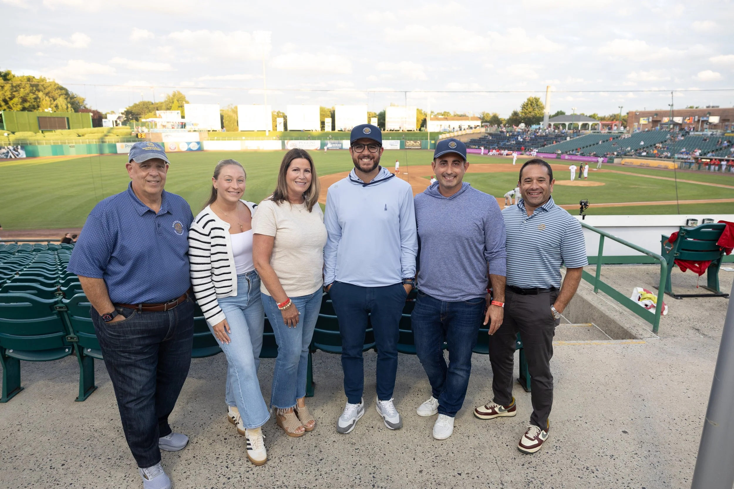 Six people are standing together in front of a baseball field during daylight. The group consists of two women and four men, all smiling. The baseball field and stands are visible in the background.