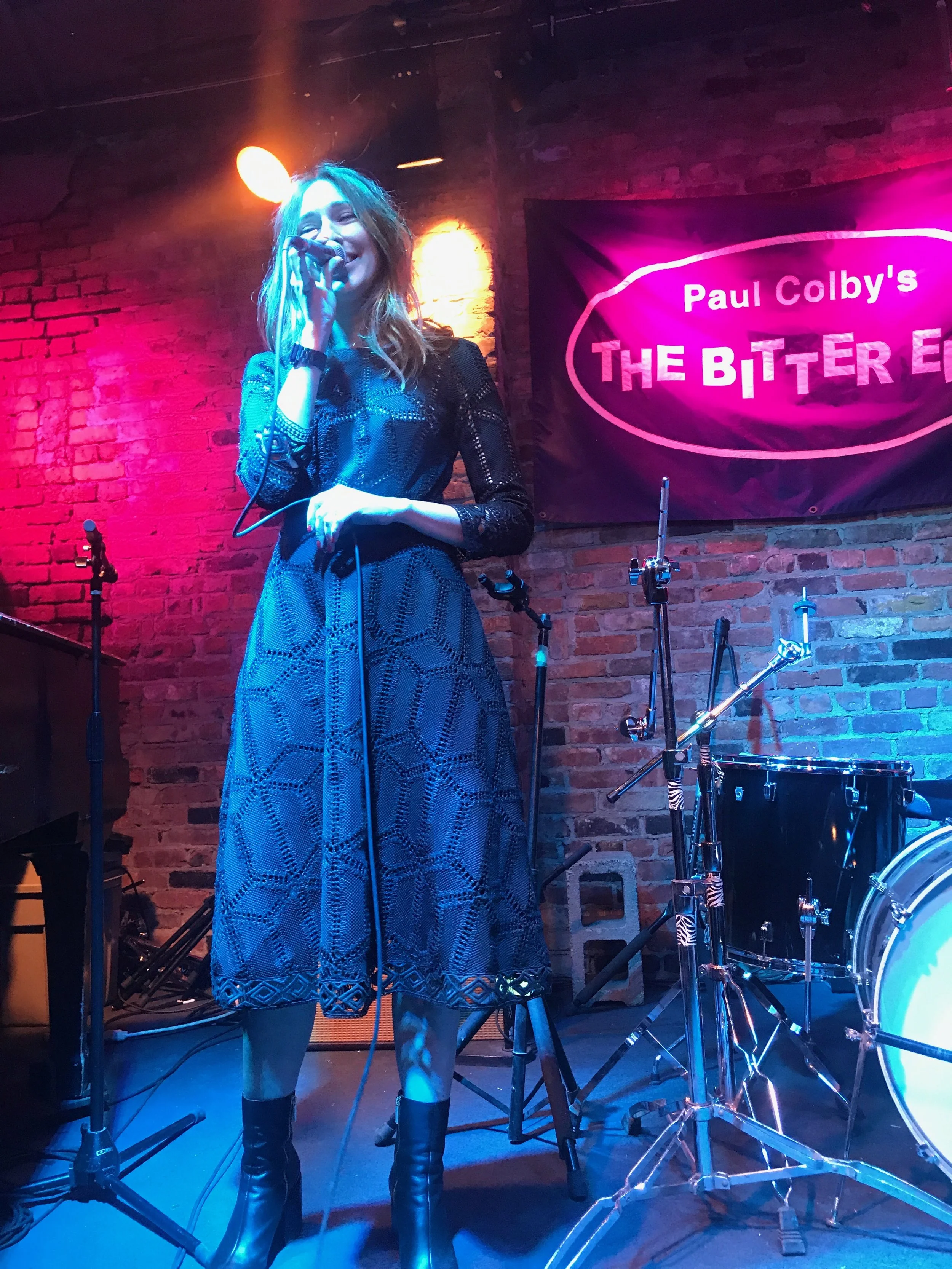 A woman with long hair singing into a microphone on stage at a jazz club, with a drum set and a black banner in the background reading 'Paul Colby's The Bitter End.'