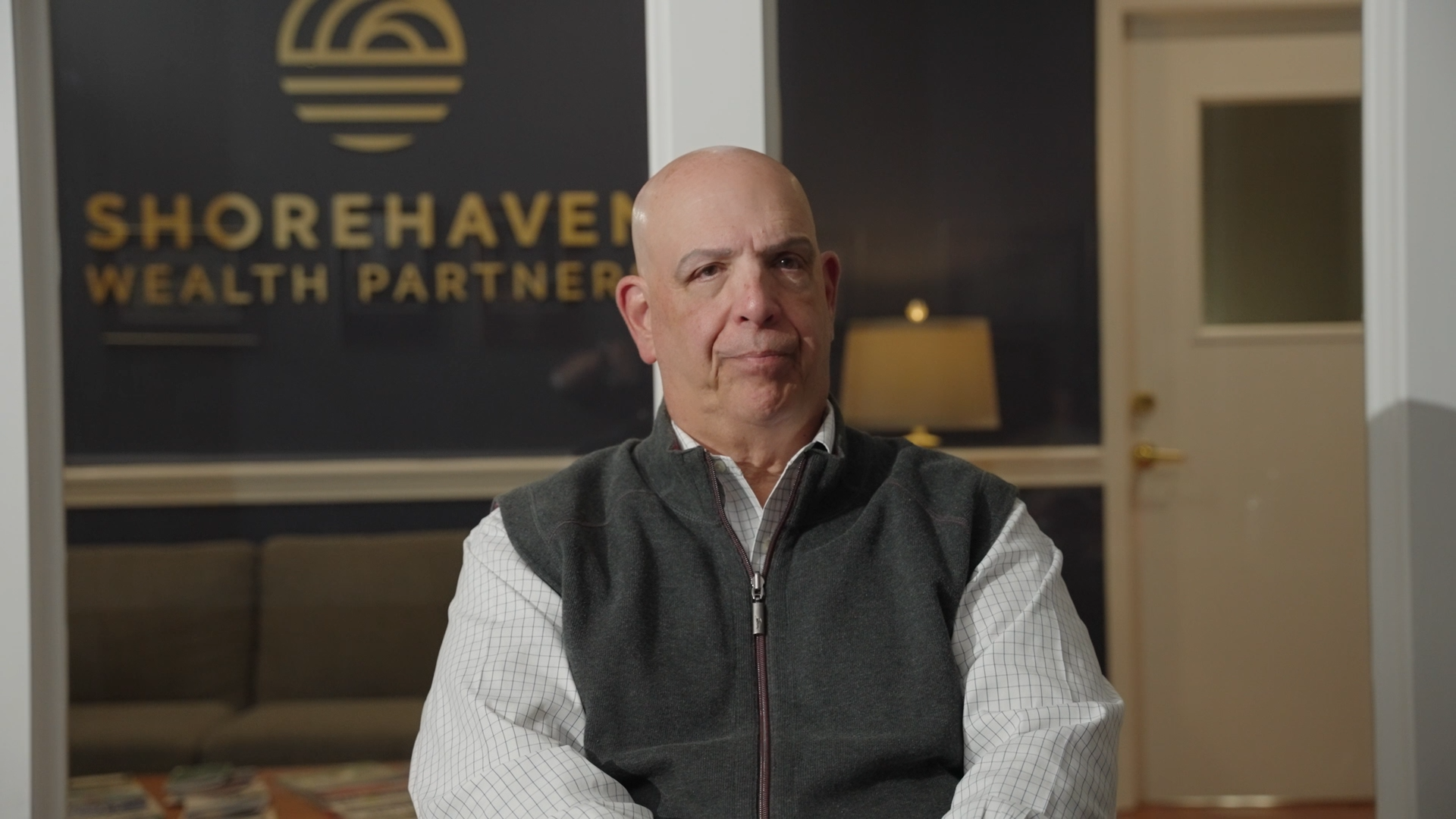 A man with a serious expression sits in front of a Shorehaven Wealth Partners sign in an office.