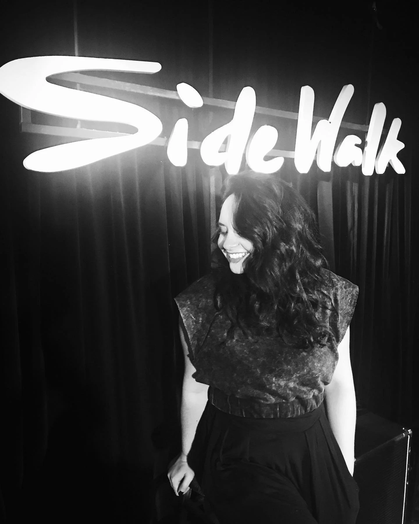A woman with curly hair smiling and looking down, standing in front of a neon sign that reads "Side Walk."