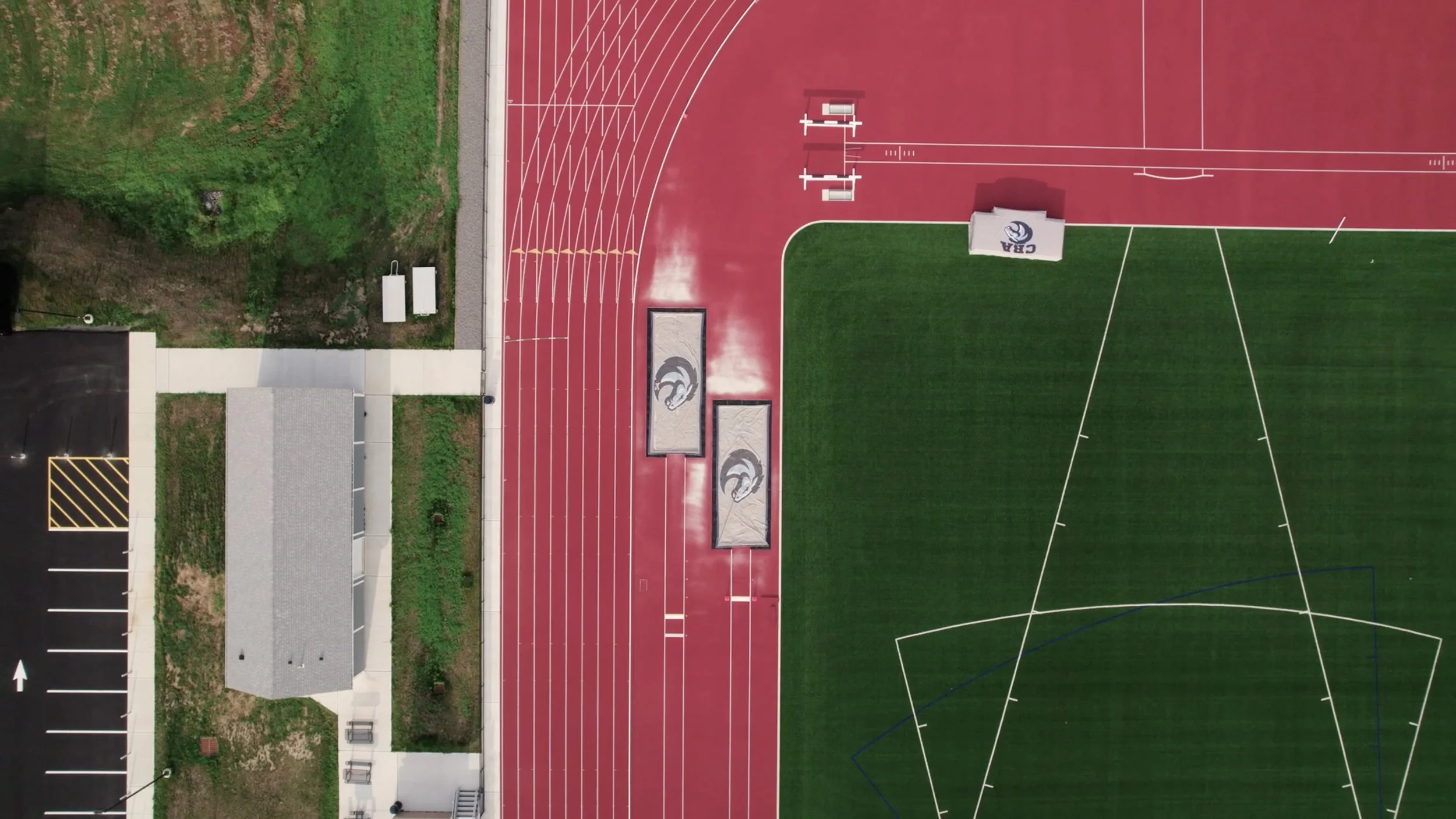An aerial view of a sports complex showing a running track, a football field, and surrounding facilities. The running track is red with white lane markings, and the football field has green grass with white lines. There is a building with a gray roof
