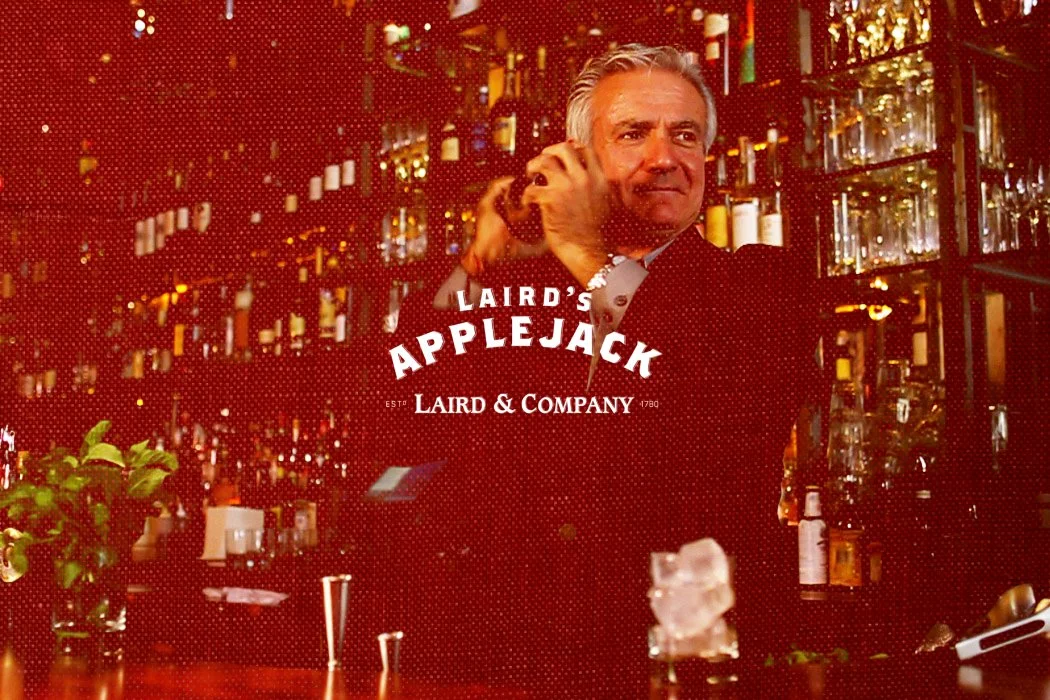 A man with gray hair wearing a dark suit and white shirt, standing behind a bar with bottles of alcohol. The bar has a warm, dim lighting ambiance. The text overlay says "Laird's Applejack, Laird & Company 1780."