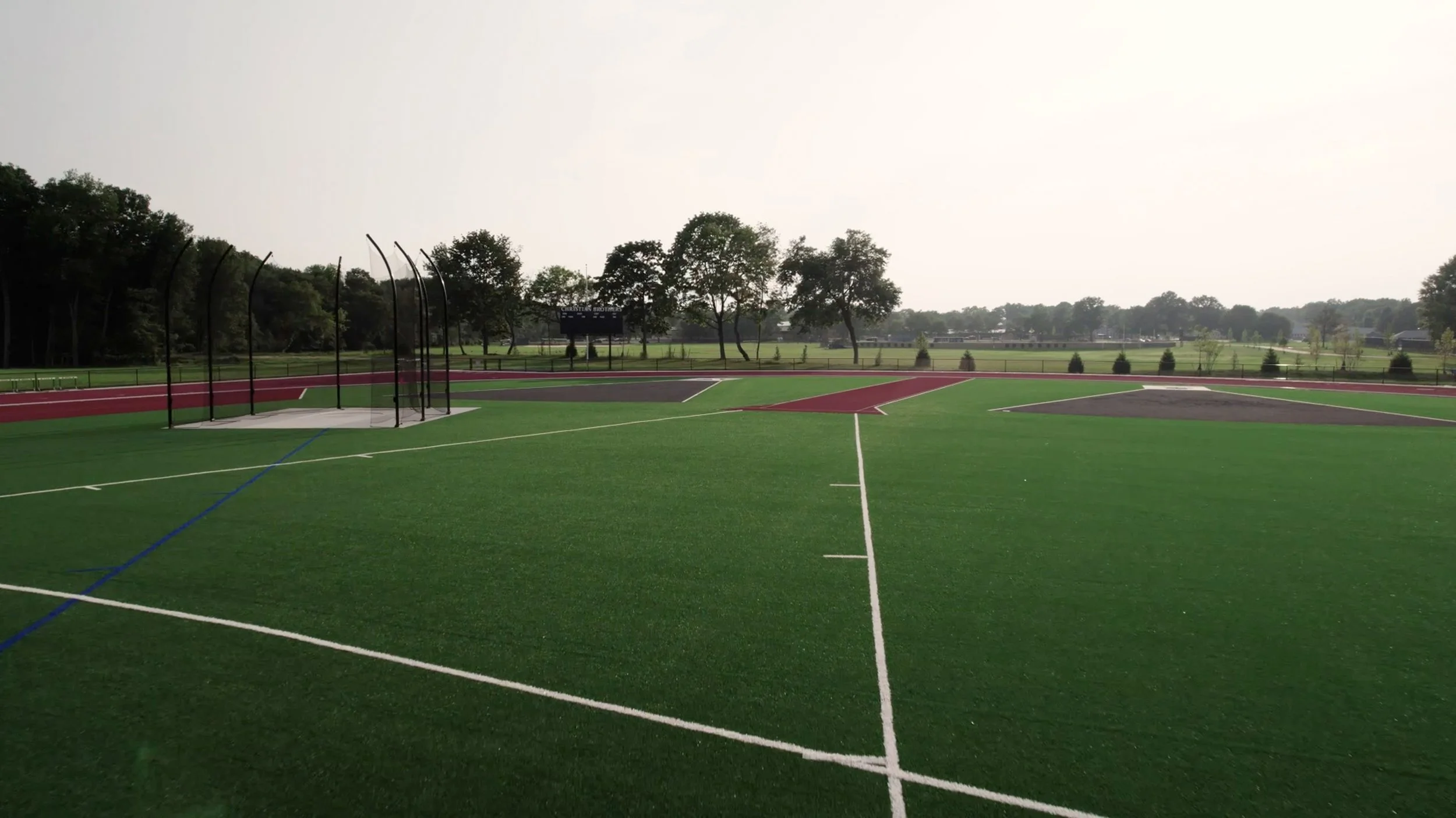 A baseball or softball field with a backstop, with a large open grassy outfield and trees in the background. The field has bright green artificial turf with painted lines.