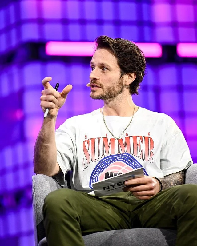 A man with brown hair and a beard sitting on a gray chair, holding a notepad in his left hand and gesturing with his right hand, in front of a purple background with pink lights, wearing a white T-shirt with a "Summer" graphic and a gold chain.