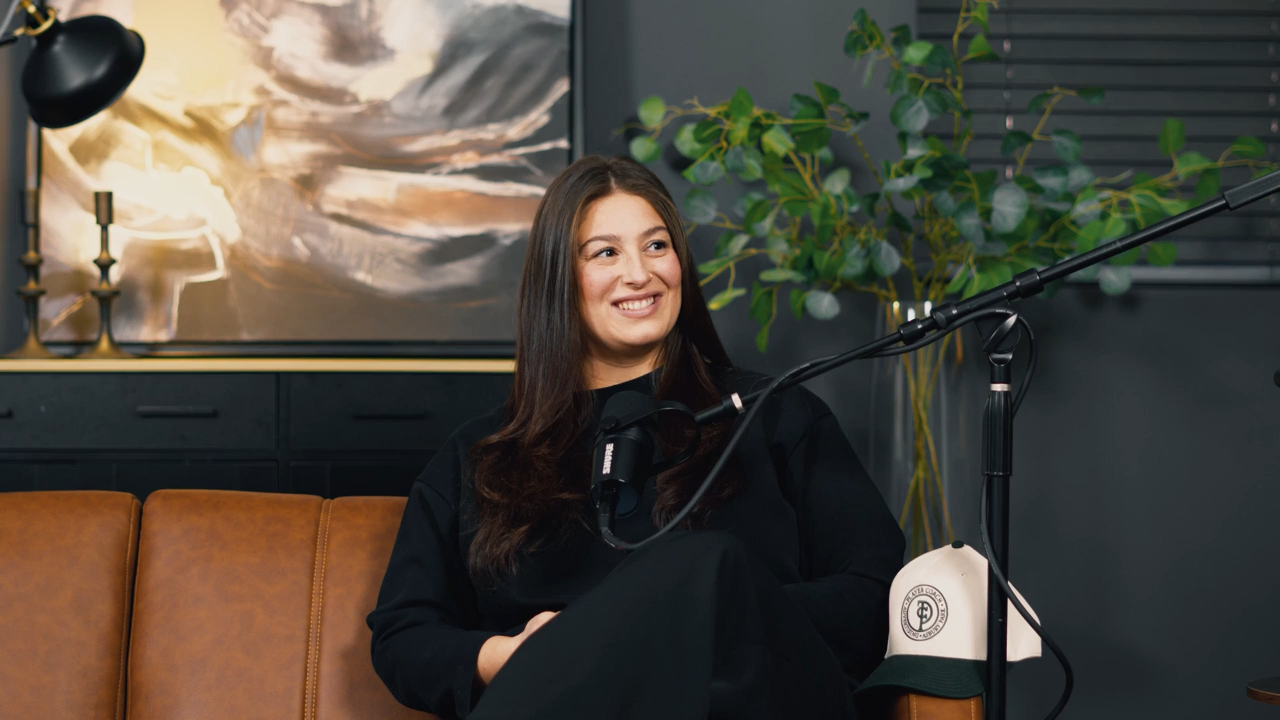 A woman with long brown hair sitting on a tan couch, smiling, during a podcast or interview session with a microphone in front of her, in a modern living room decorated with a large painting, a black cabinet, a table lamp, and a potted plant.